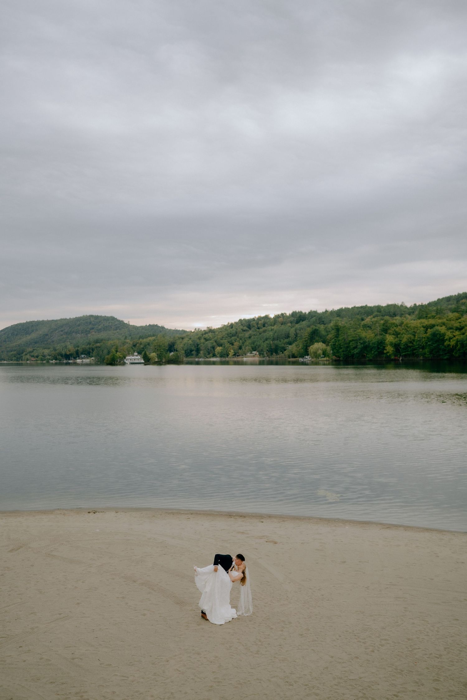 Couple in wedding attire standing on sandy beach by calm lake with forested hills and cloudy sky backdrop.