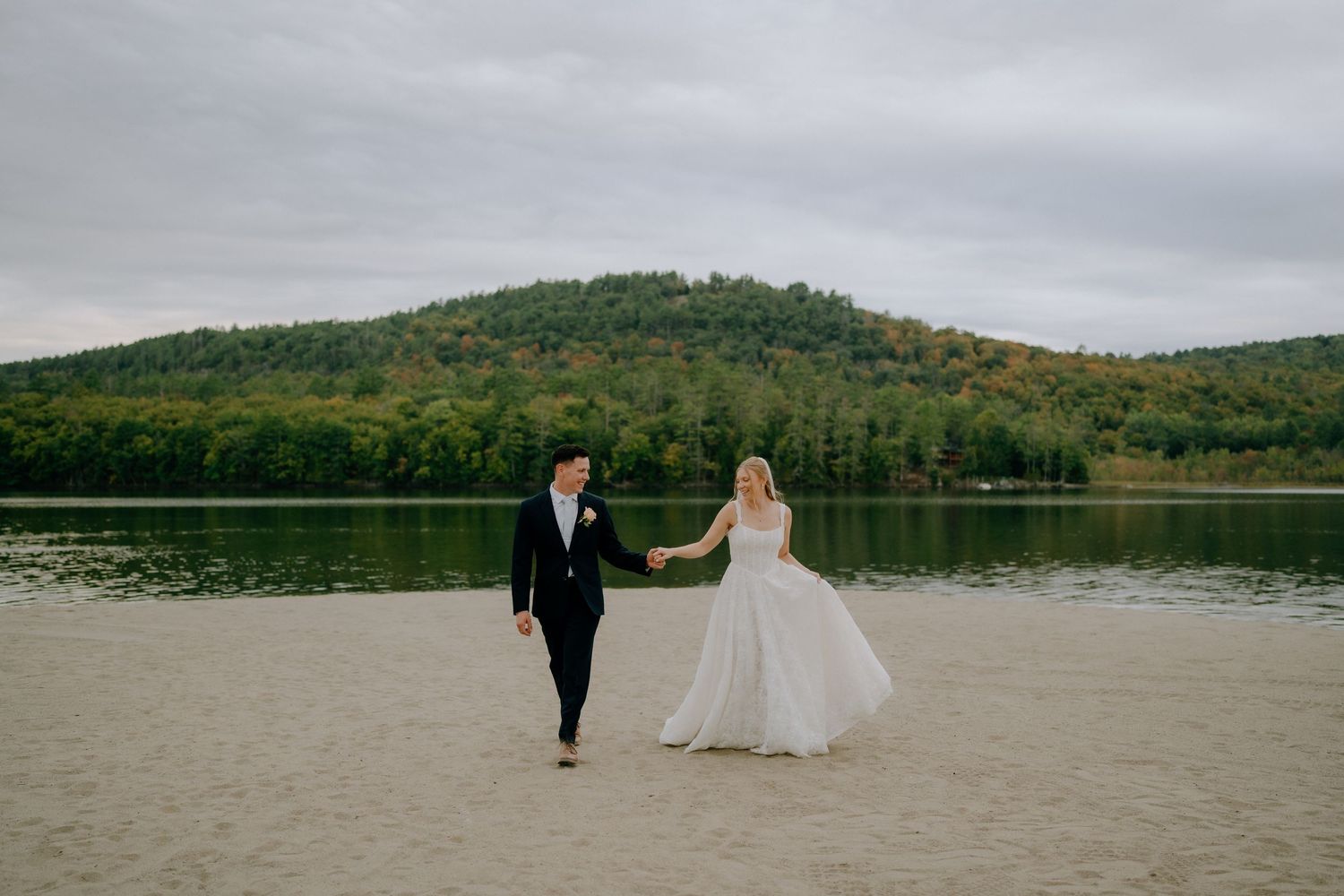 Bride and groom walk hand in hand on sandy beach beside a serene lake with lush green hills in background.