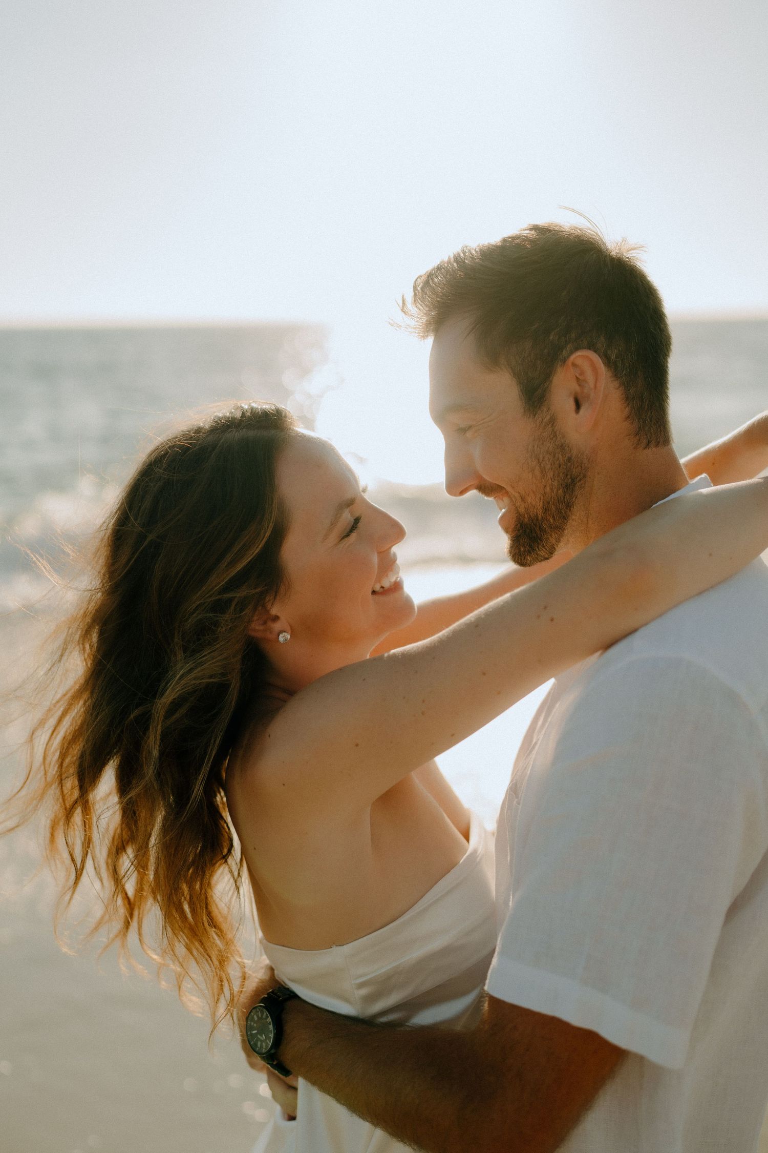A couple embraces and smiles at each other on a sunny beach, with golden backlight and wind-swept hair.