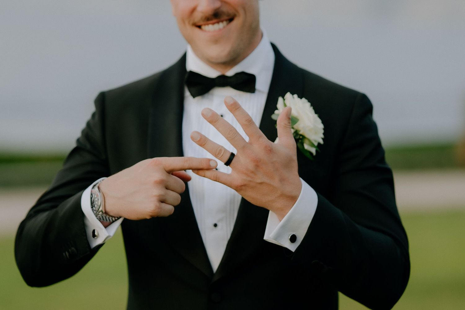 Groom in black tuxedo with bow tie pointing to wedding ring on finger, white boutonniere on lapel.
