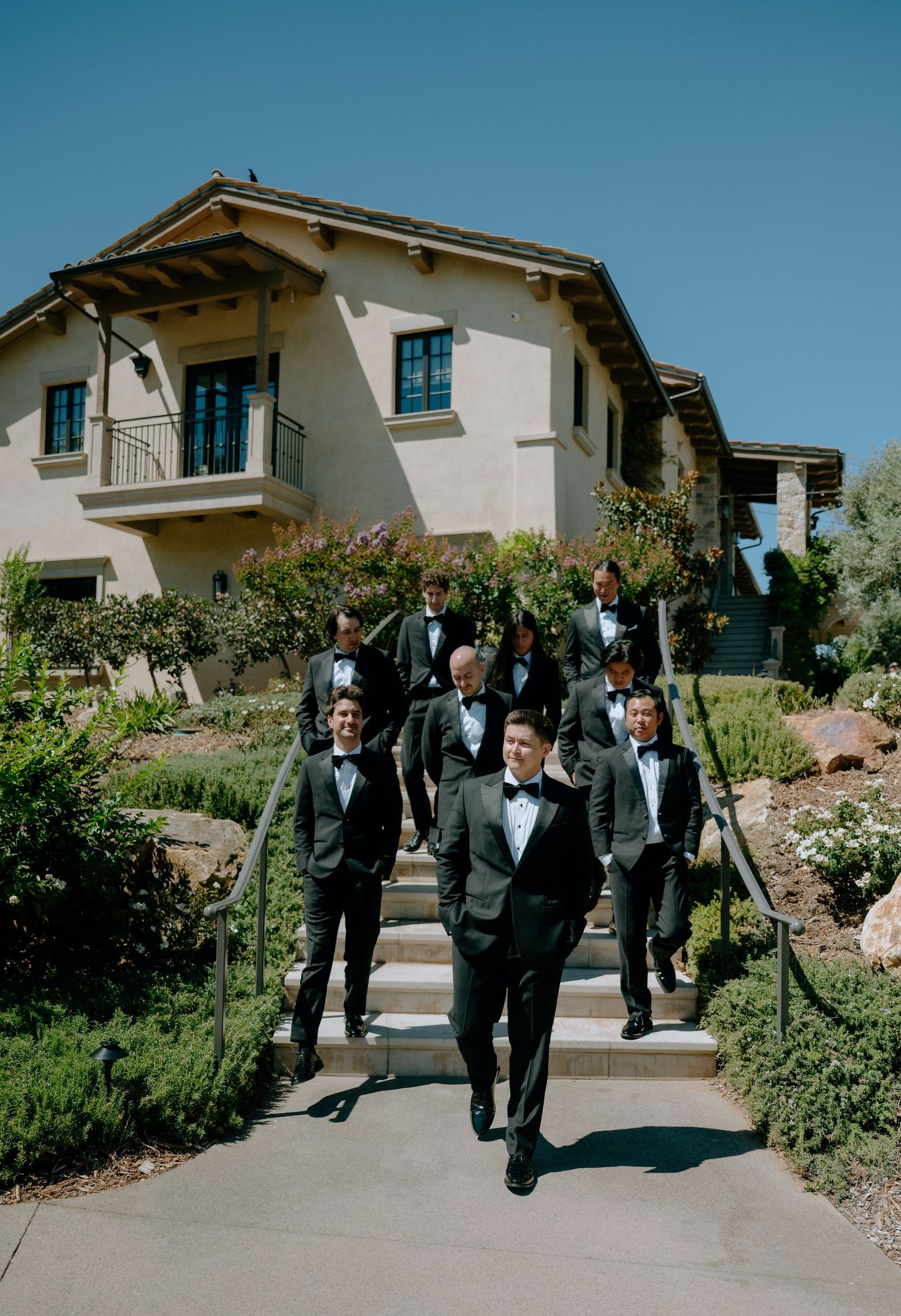 Groom and groomsmen in black tuxedos descending outdoor steps at an elegant wedding venue.