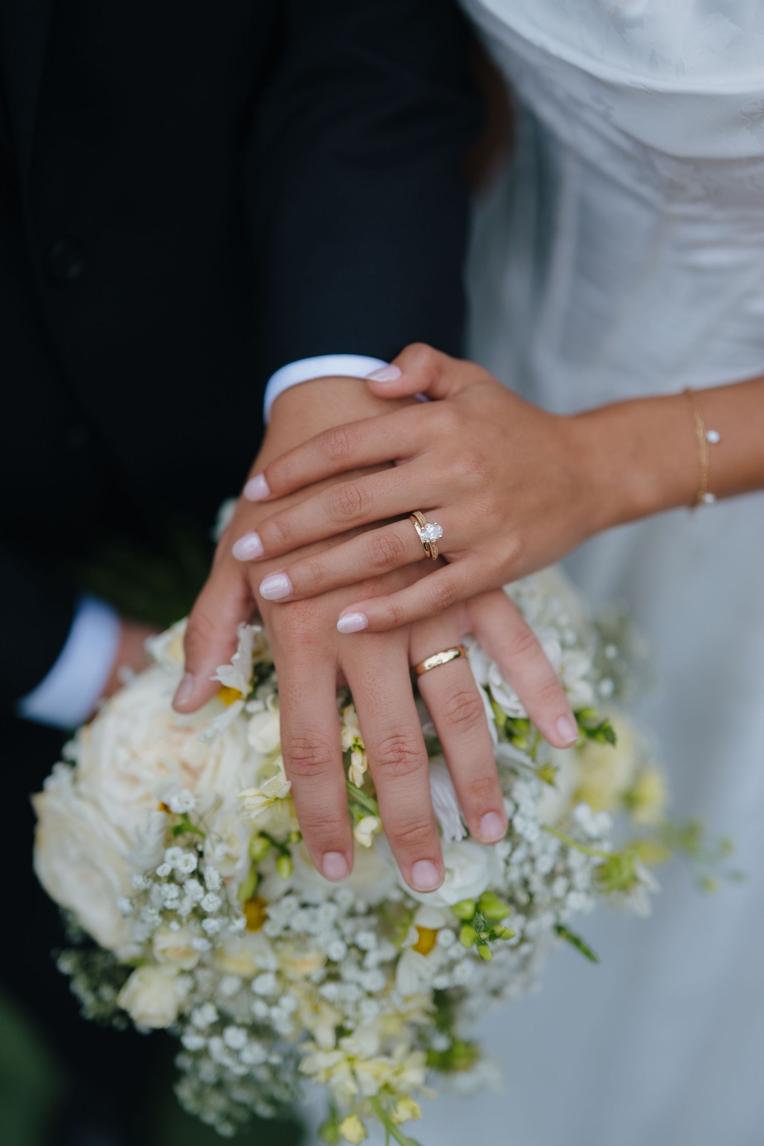 Close-up of newlyweds hands displaying wedding rings resting on a white bridal bouquet.