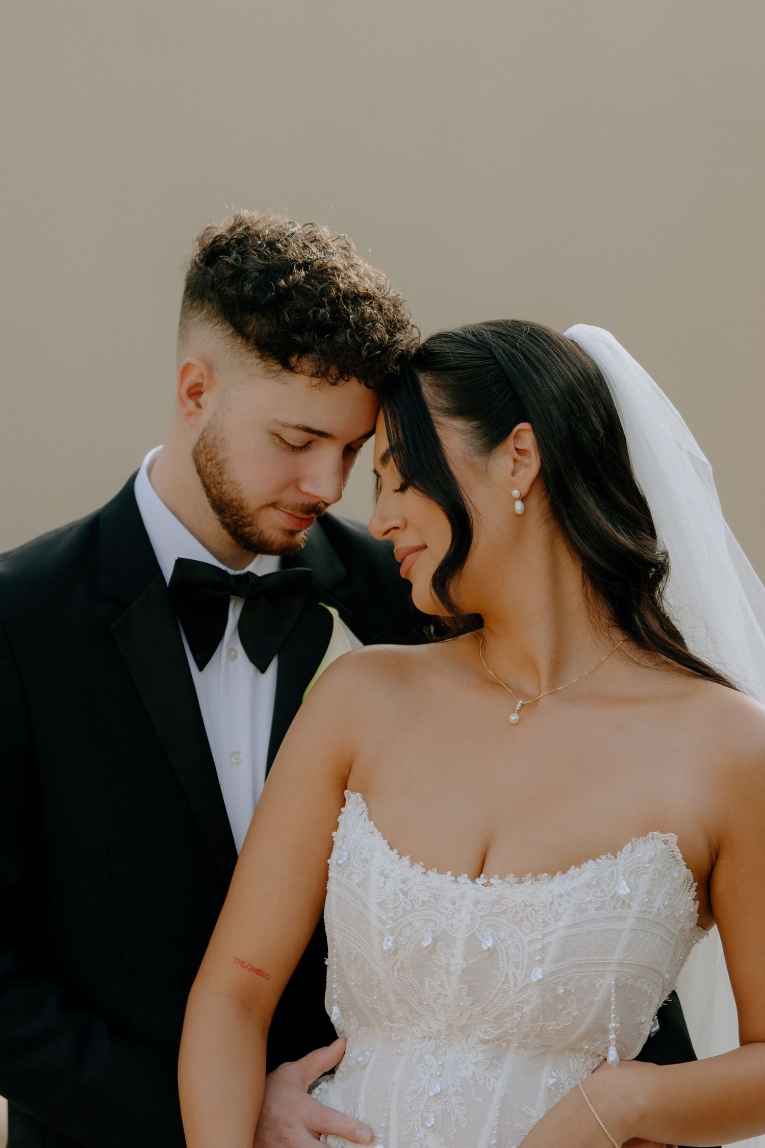 A bride and groom share an intimate forehead touch, she in a white strapless gown with veil, he in a black tuxedo.