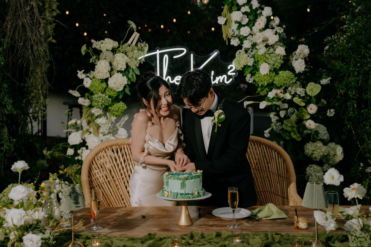 Couple cutting a green wedding cake at a floral-decorated reception table with neon sign backdrop.