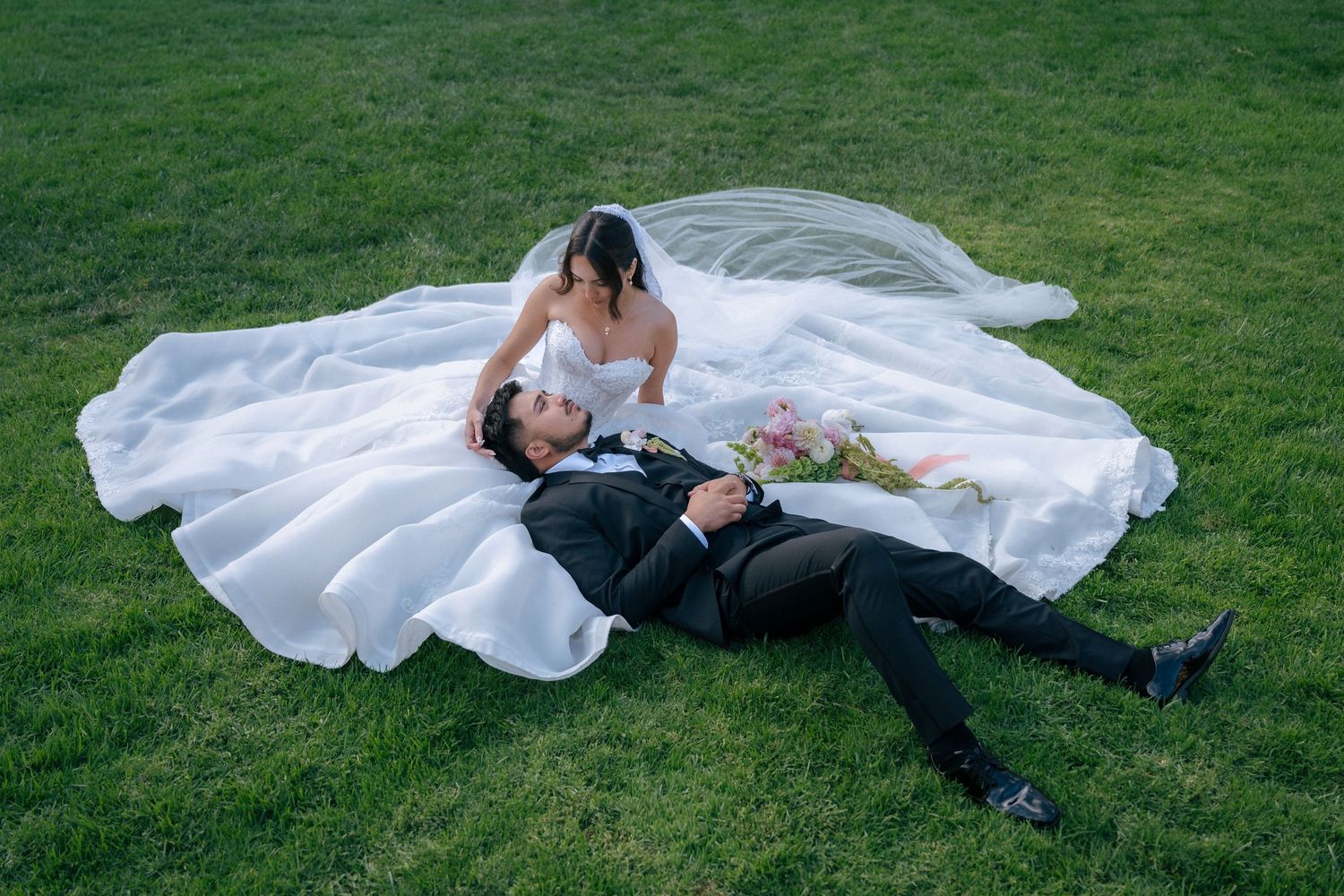 Bride in white gown sits beside groom lying on green grass, her veil spread wide, holding a bouquet.