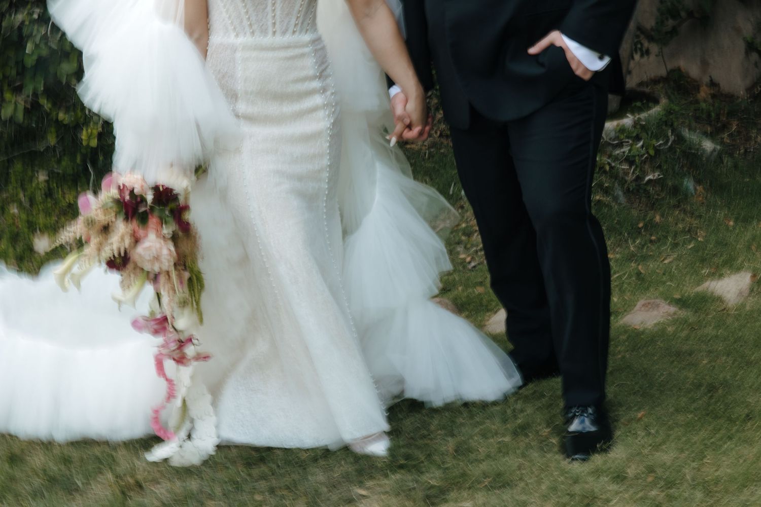 Close-up of bride in lace gown holding a cascading pink floral bouquet while walking with groom.