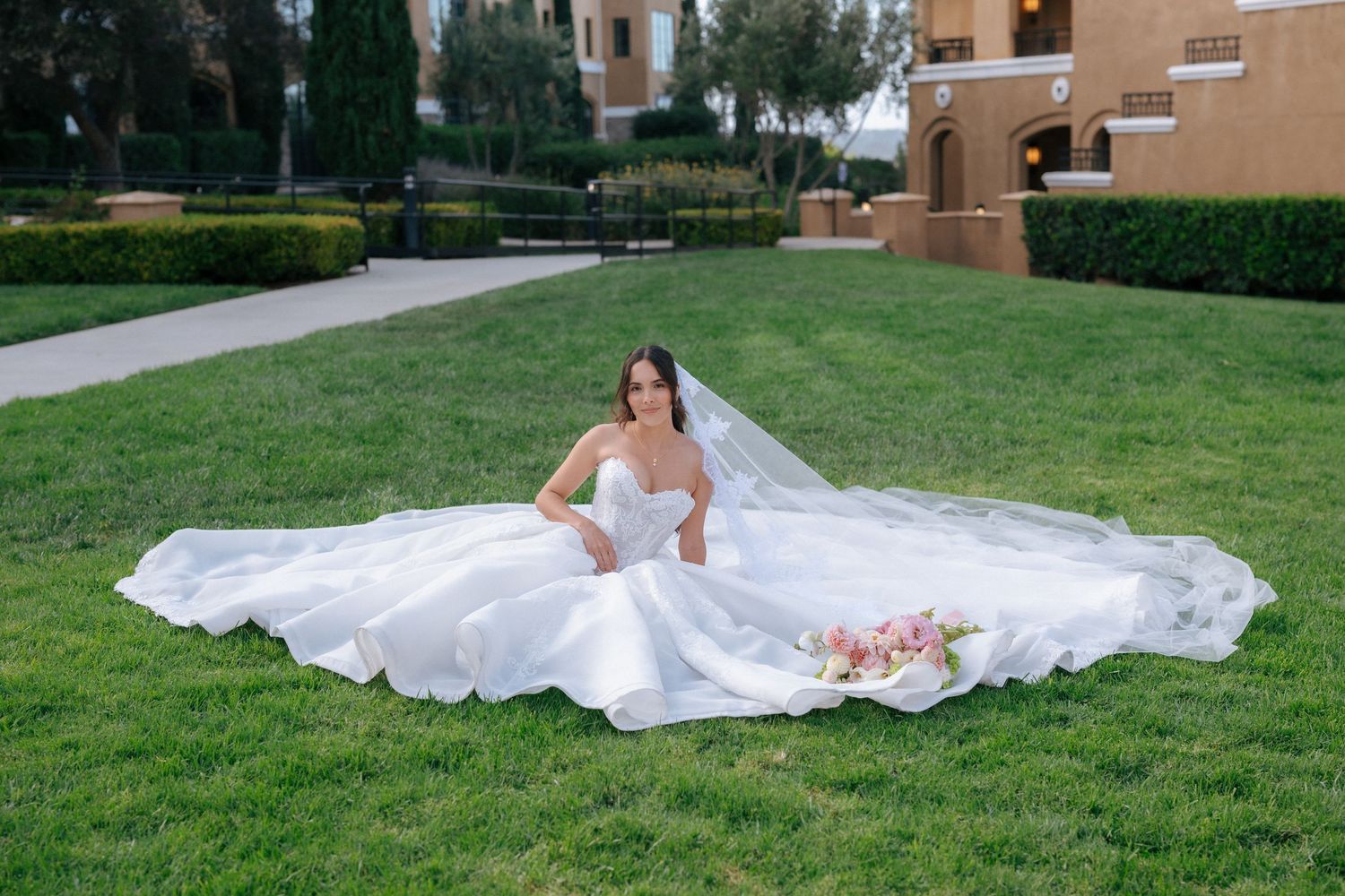 Bride in a stunning white ball gown with long veil sits on green grass, holding a pink bouquet at an outdoor venue.