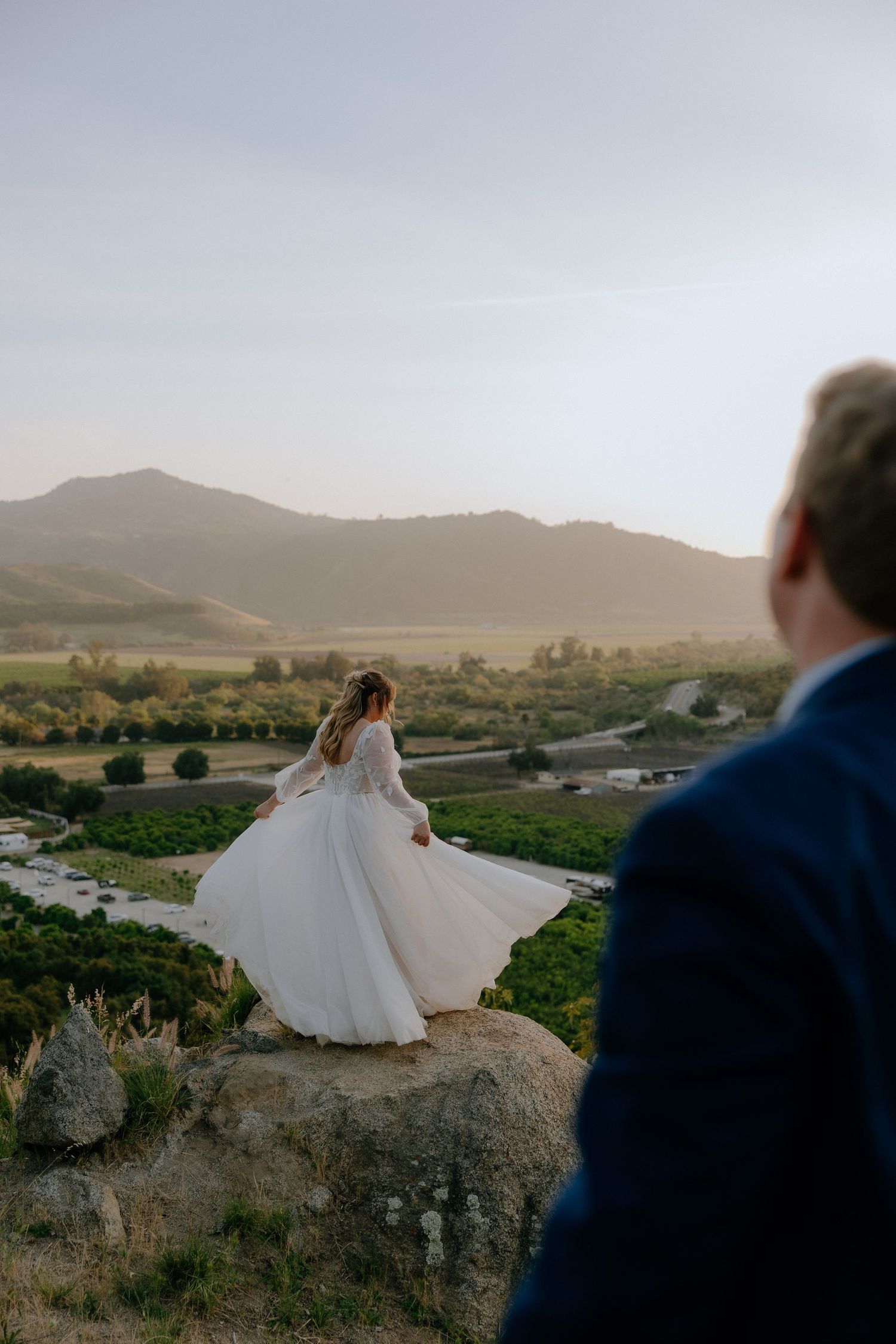Bride in flowing white gown twirling on a rock with scenic vineyard and mountain views at golden hour.