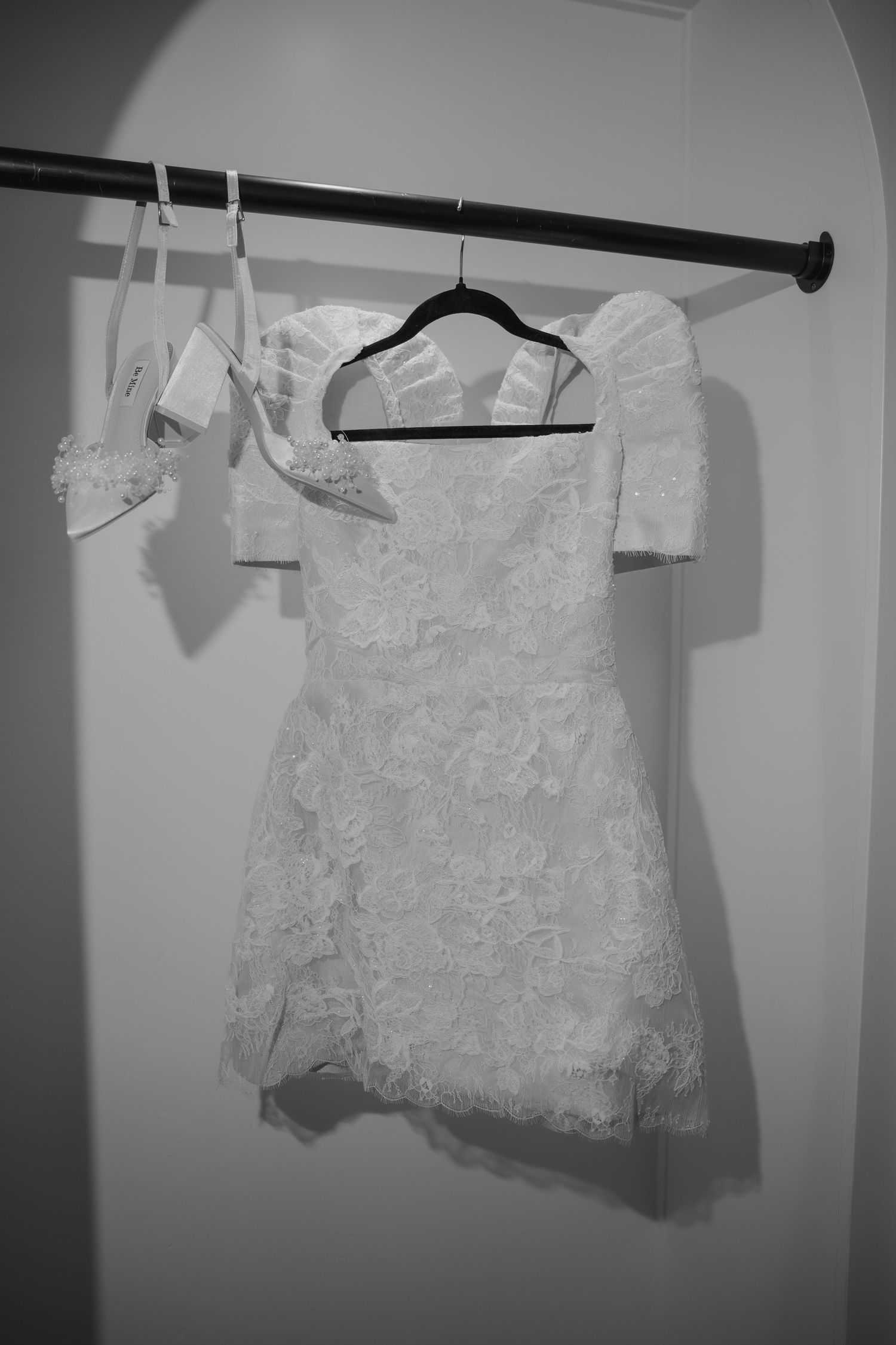 Black and white photo of a lace off-shoulder mini dress on a hanger with white heels on a closet rod.
