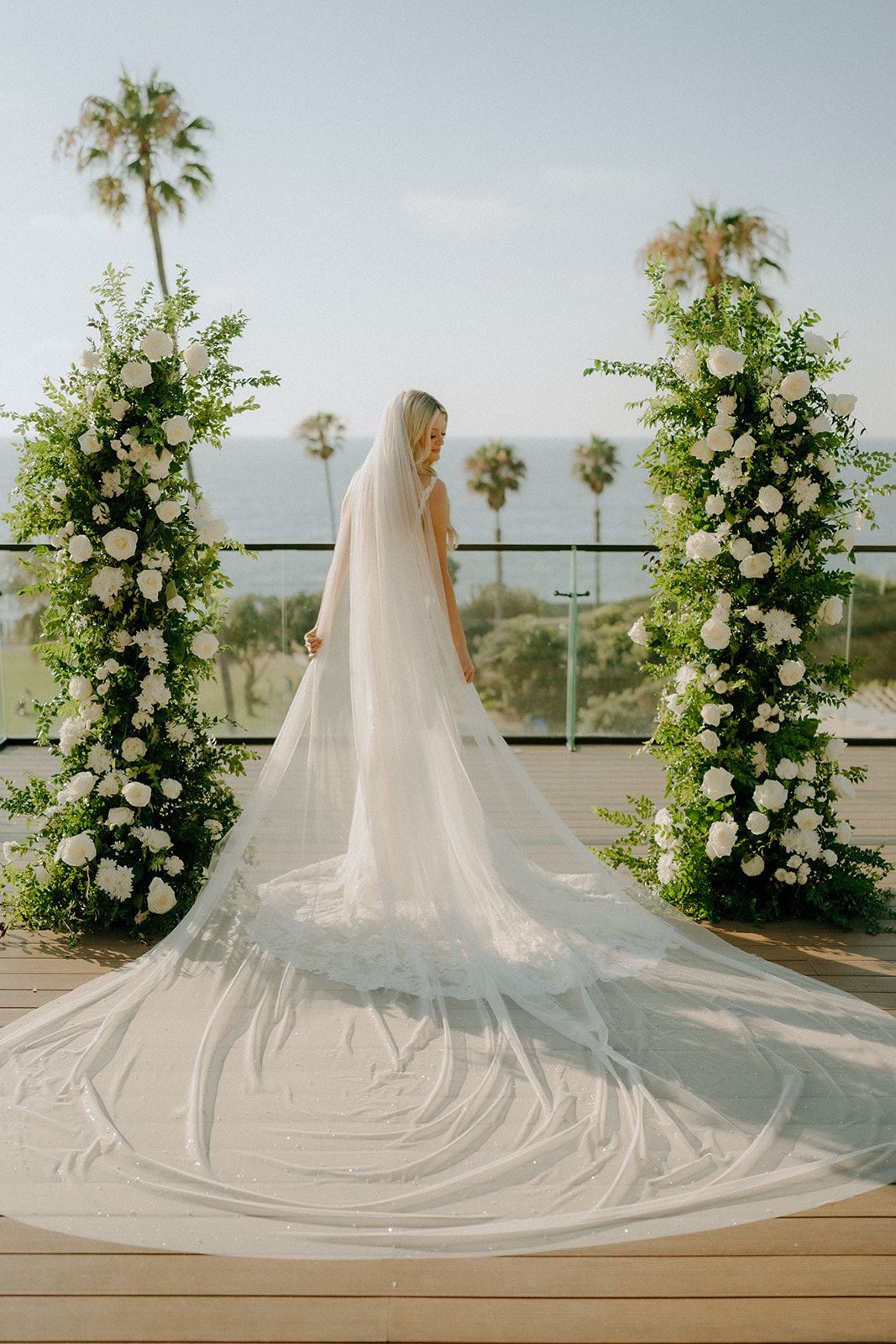 Bride in white gown and cathedral veil stands between floral columns with white roses and greenery on an outdoor terrace.