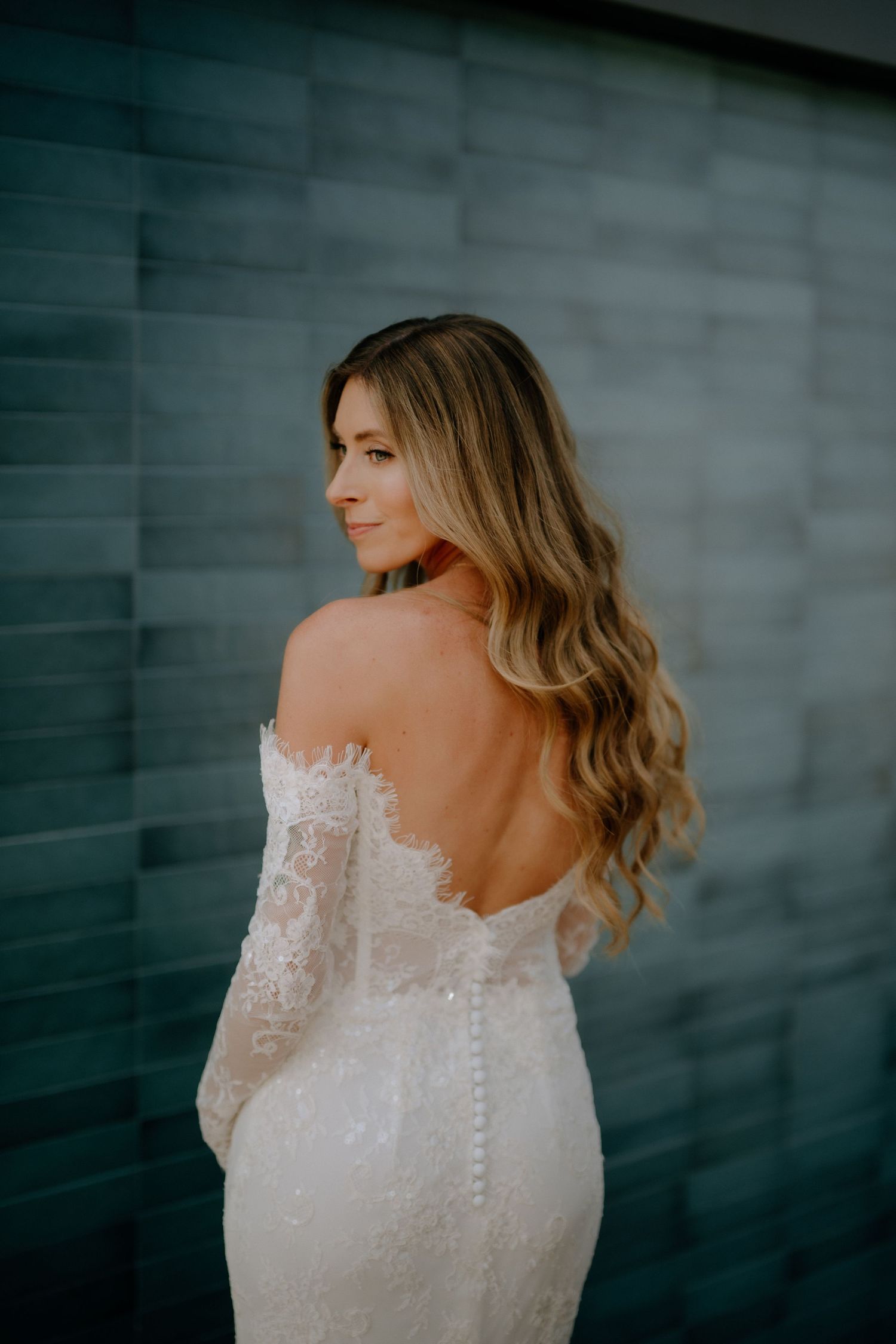 Bride in off-shoulder lace wedding gown with open back, showcasing wavy blonde hair against a tiled wall.