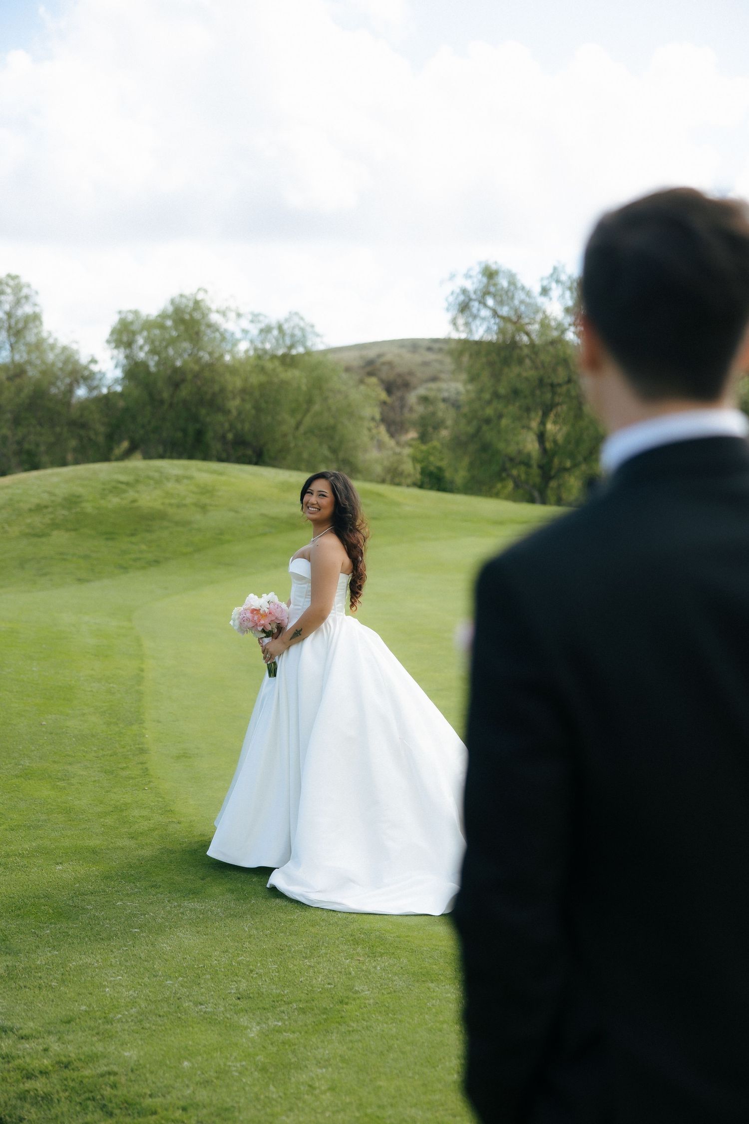 Bride in white ballgown holding pink bouquet smiles at groom in black suit on lush green golf course.