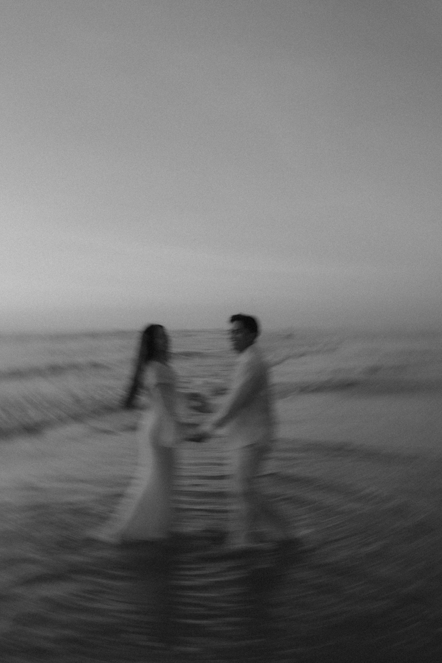 Black and white photo of a couple holding hands in ocean waves at dusk, wearing wedding attire.