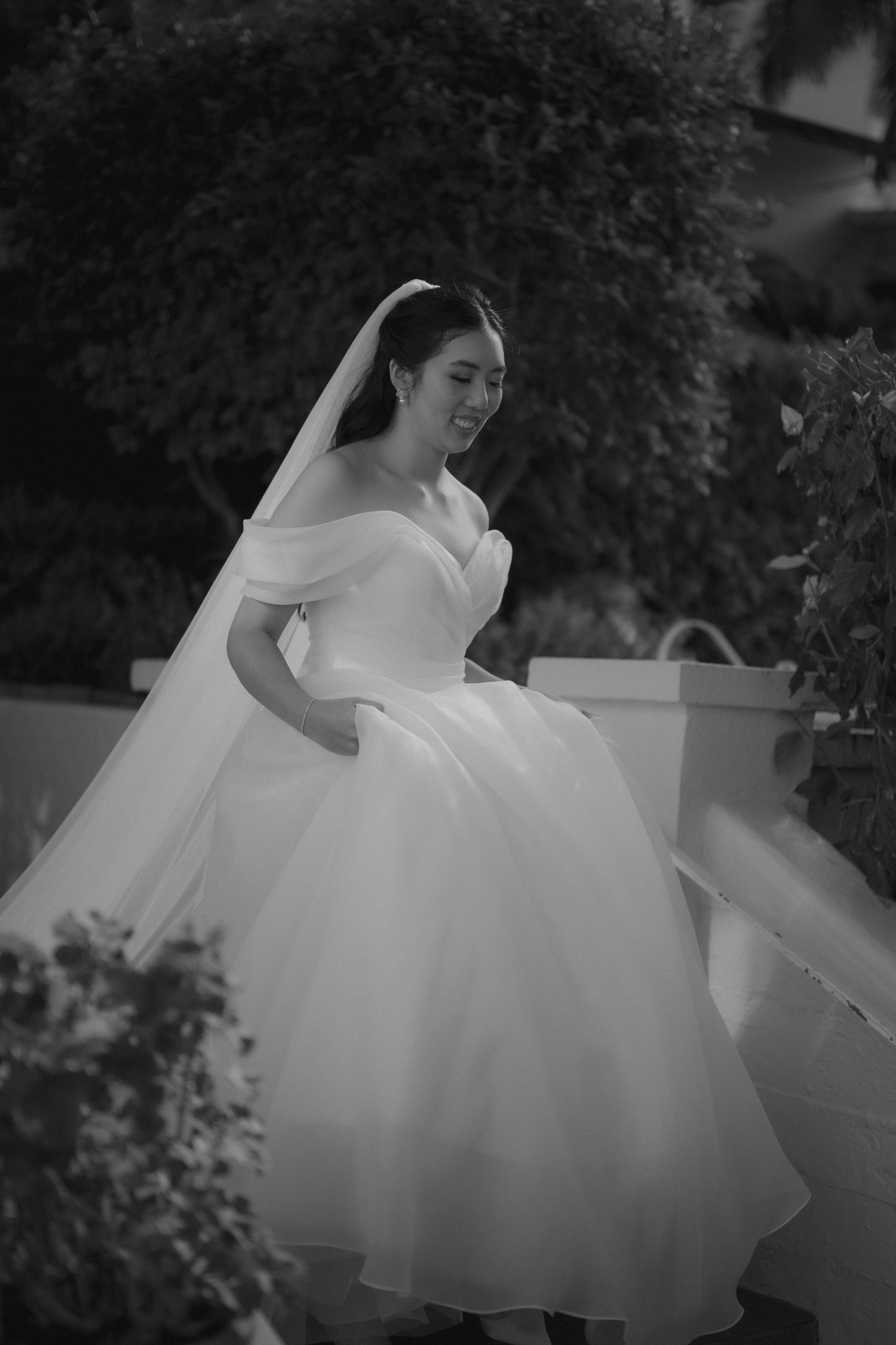 Black and white photo of a bride in an off-shoulder ball gown with veil, smiling outdoors among lush greenery.