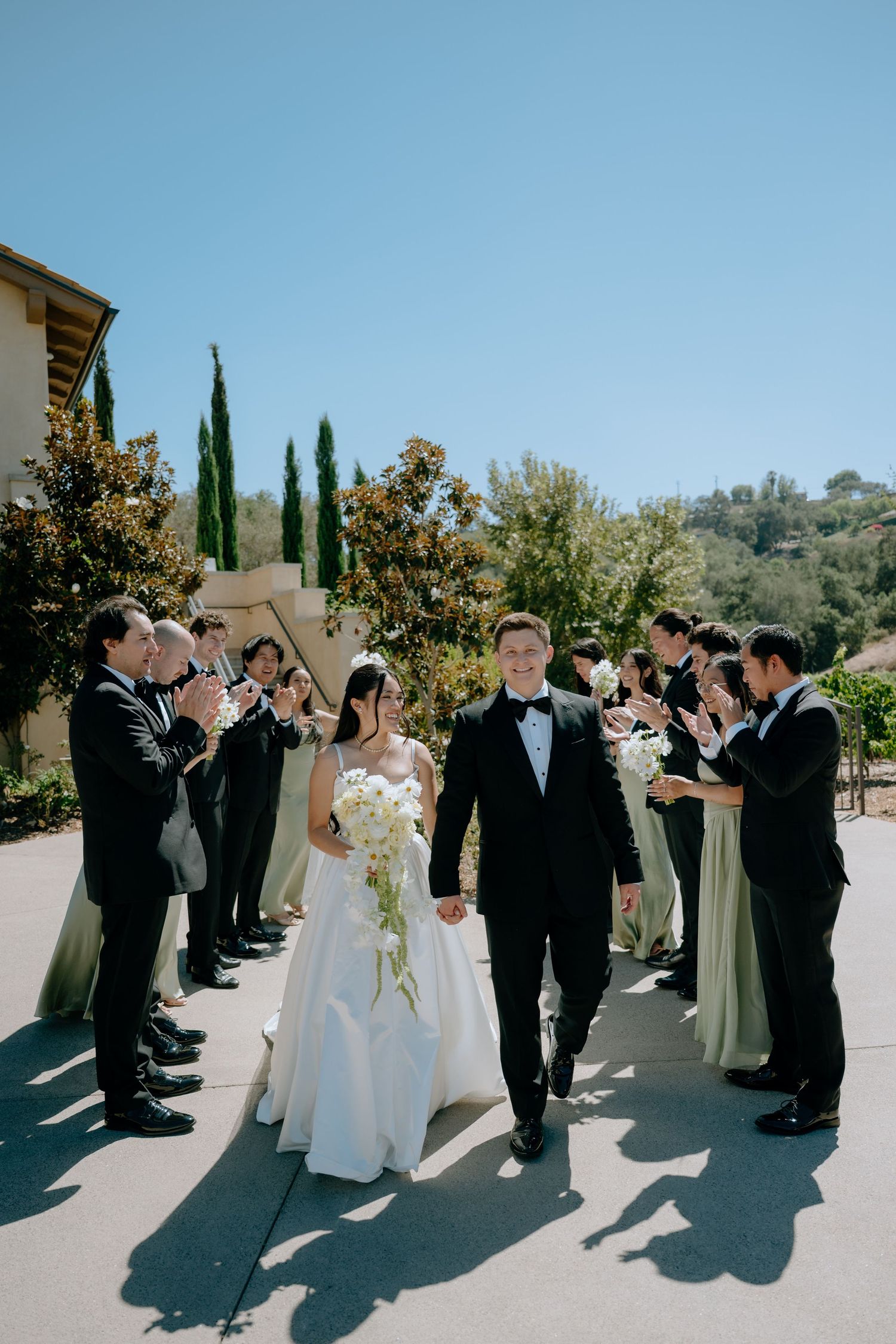 Bride and groom walk through cheering wedding party outdoors at a sunny vineyard venue, bride holding white cascading bouquet.
