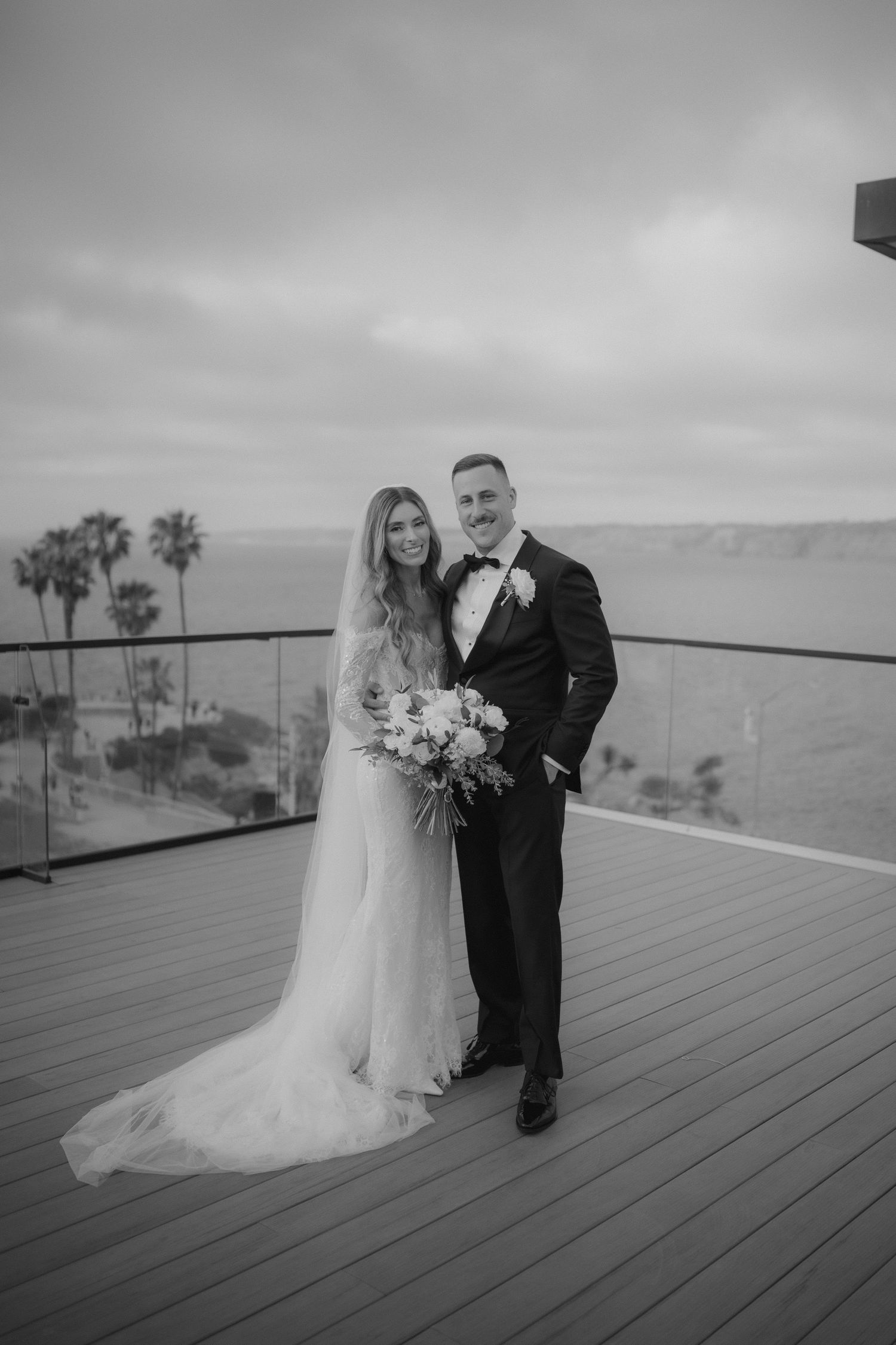 Black and white photo of a bride and groom posing on a rooftop deck with ocean views and palm trees.