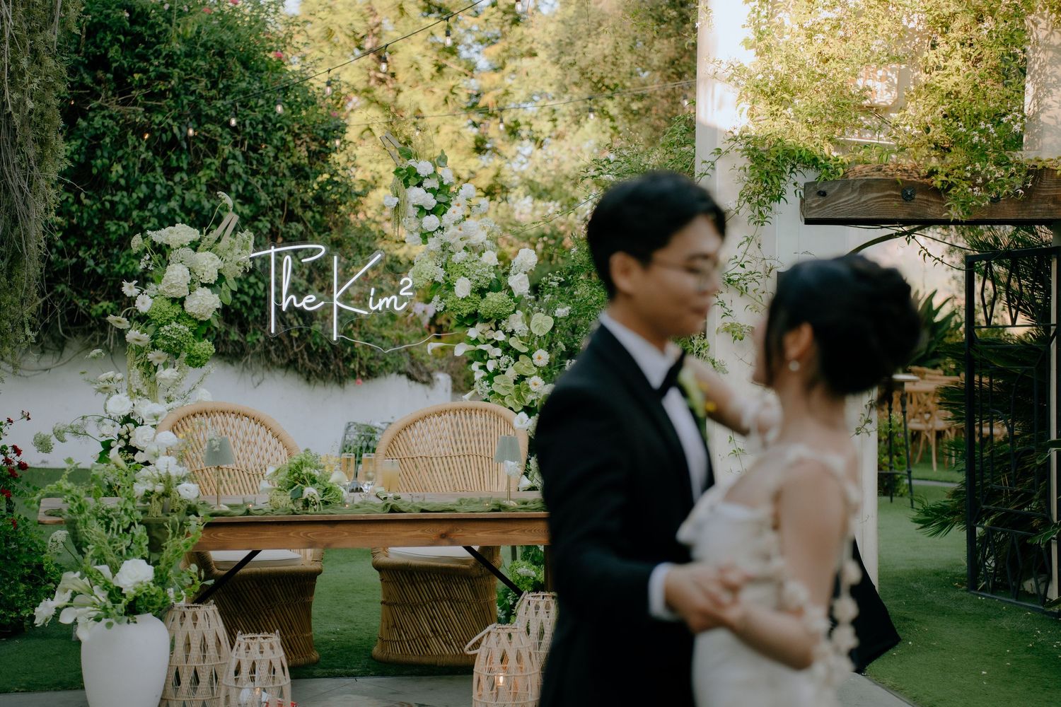 Couple embracing at elegant outdoor wedding reception with green floral backdrop and The Kims sign.