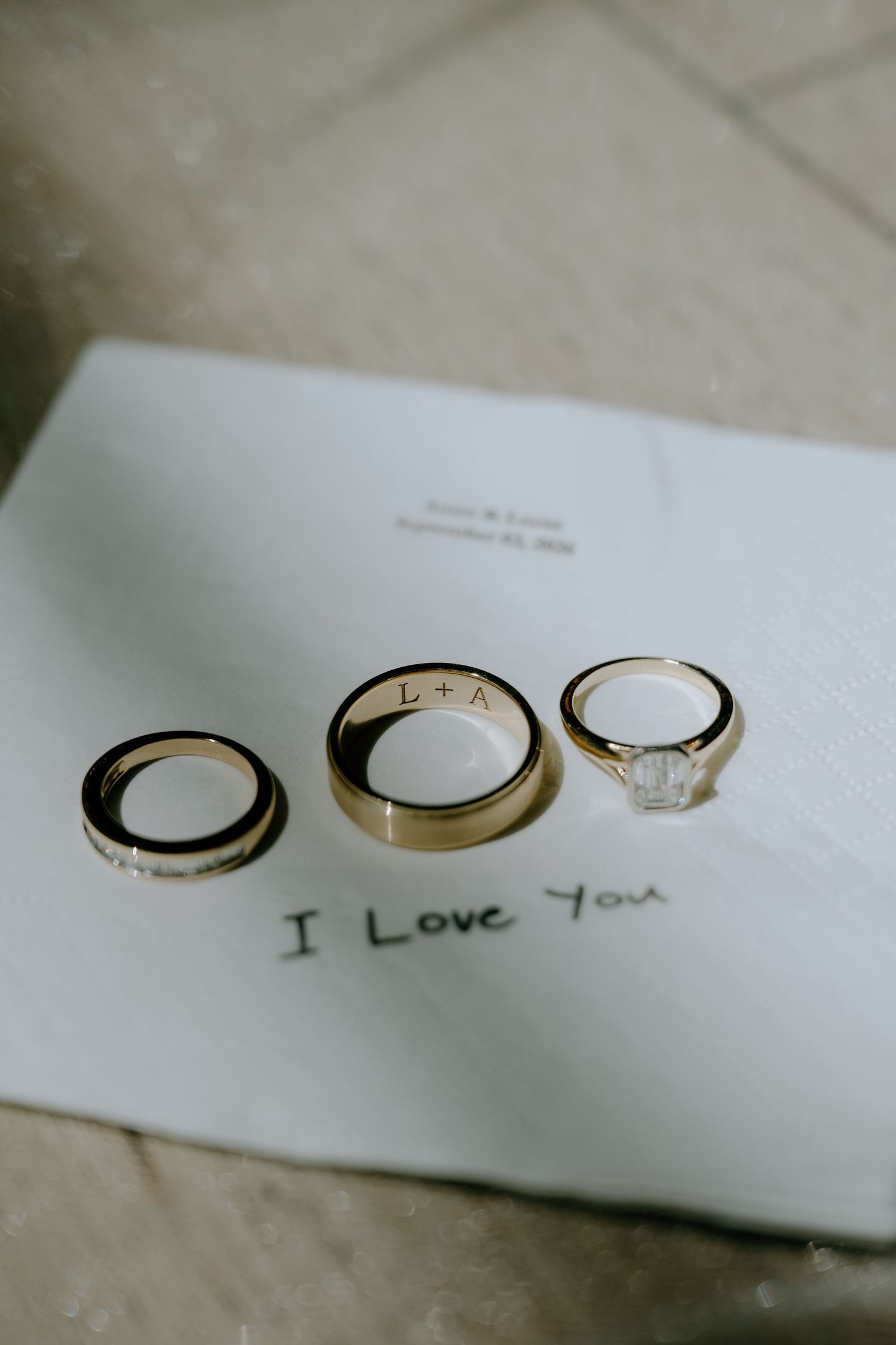 Three gold wedding rings on a love note reading I Love You, with engraved initials L+A on the center band.