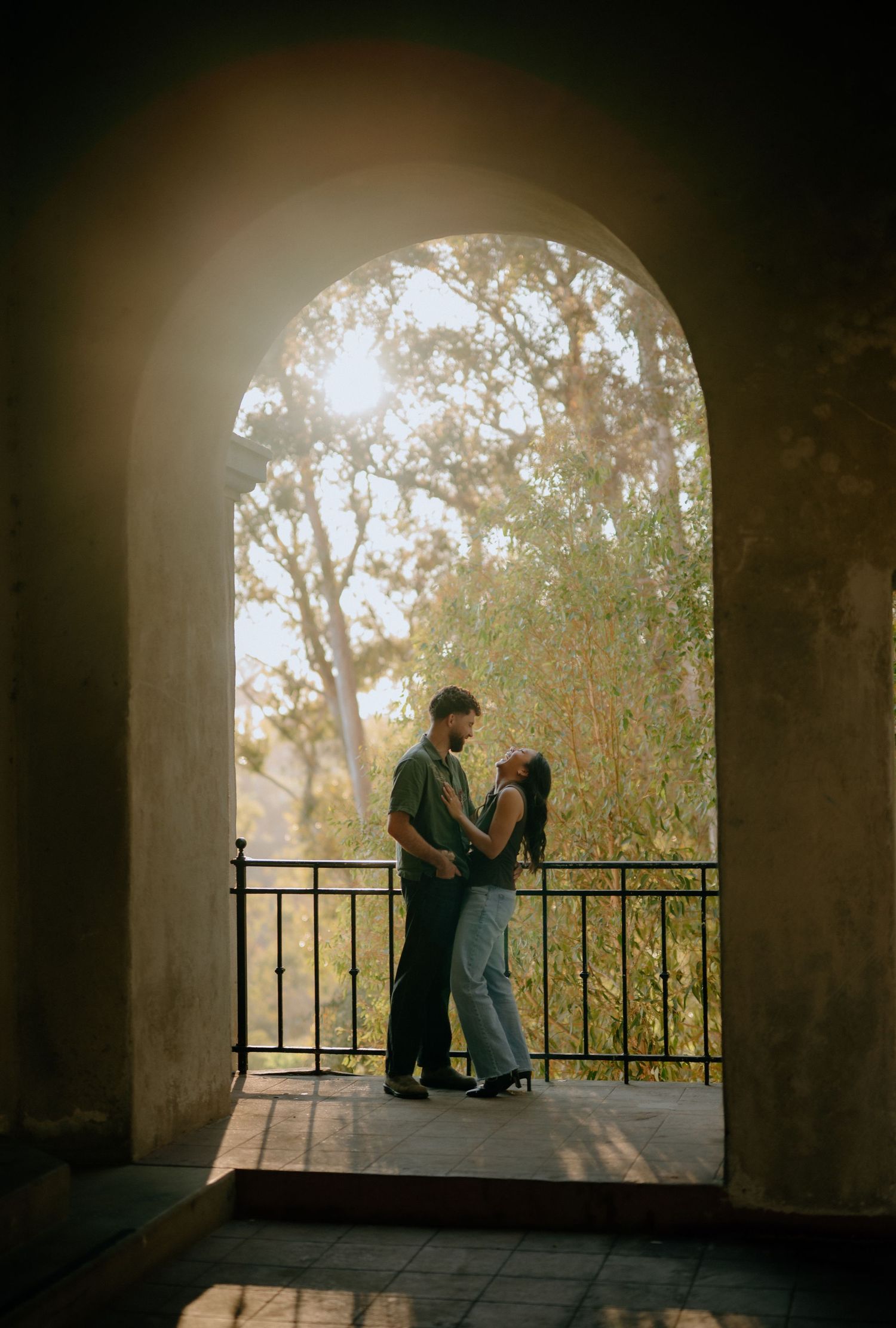 Couple embracing under a sunlit stone arch with lush trees in the background.