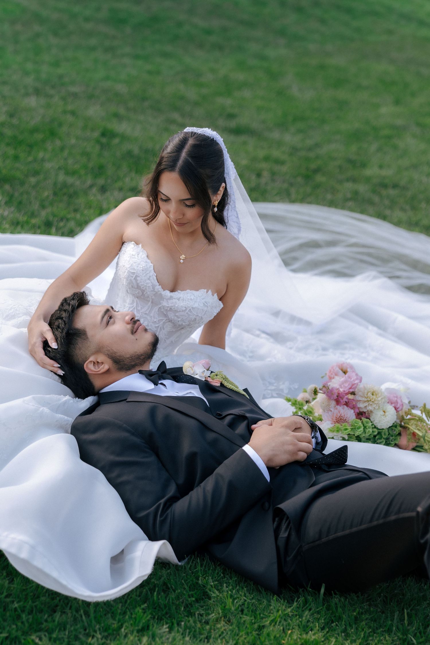 Bride in white lace gown sits on grass while groom in black tuxedo rests his head in her lap outdoors.
