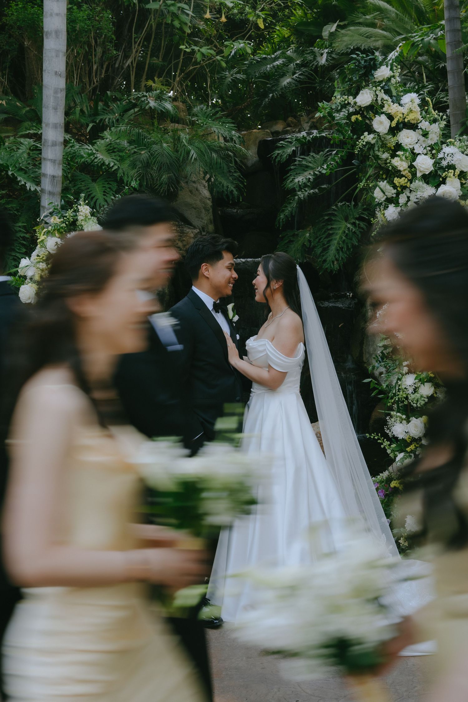 Bride and groom share an intimate moment at a lush outdoor wedding ceremony, with blurred bridesmaids in gold dresses in foreground.