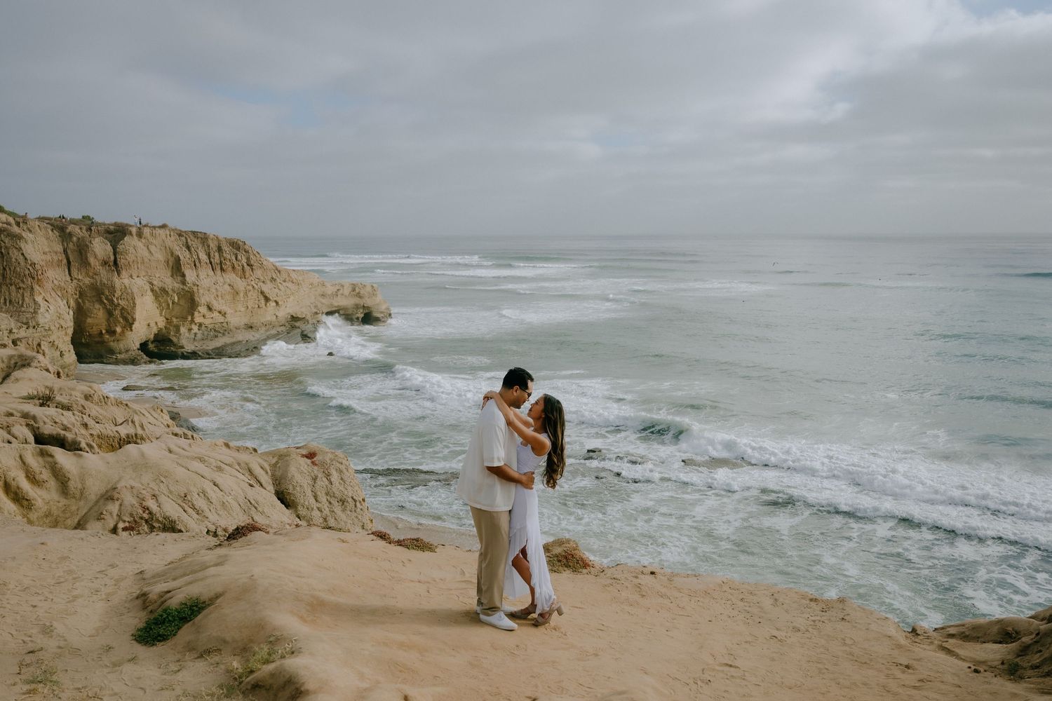 Couple embracing on a rocky coastal cliff overlooking ocean waves under an overcast sky.