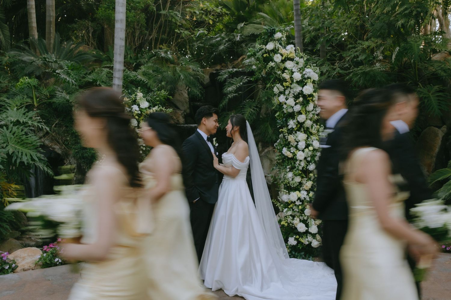 Bride and groom share a romantic moment at floral arch altar while bridesmaids in gold dresses and groomsmen walk past.