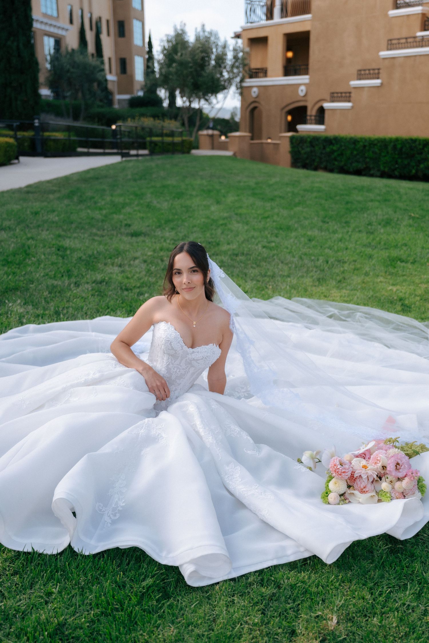 Bride in strapless ball gown sits on green grass with floral bouquet, veil flowing, at elegant outdoor venue.