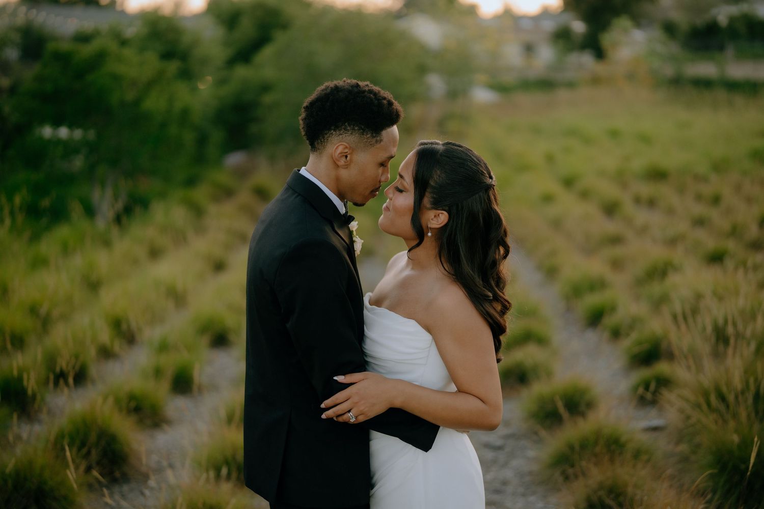 Bride and groom share a romantic moment in a lush green garden at golden hour, nearly kissing in wedding attire.