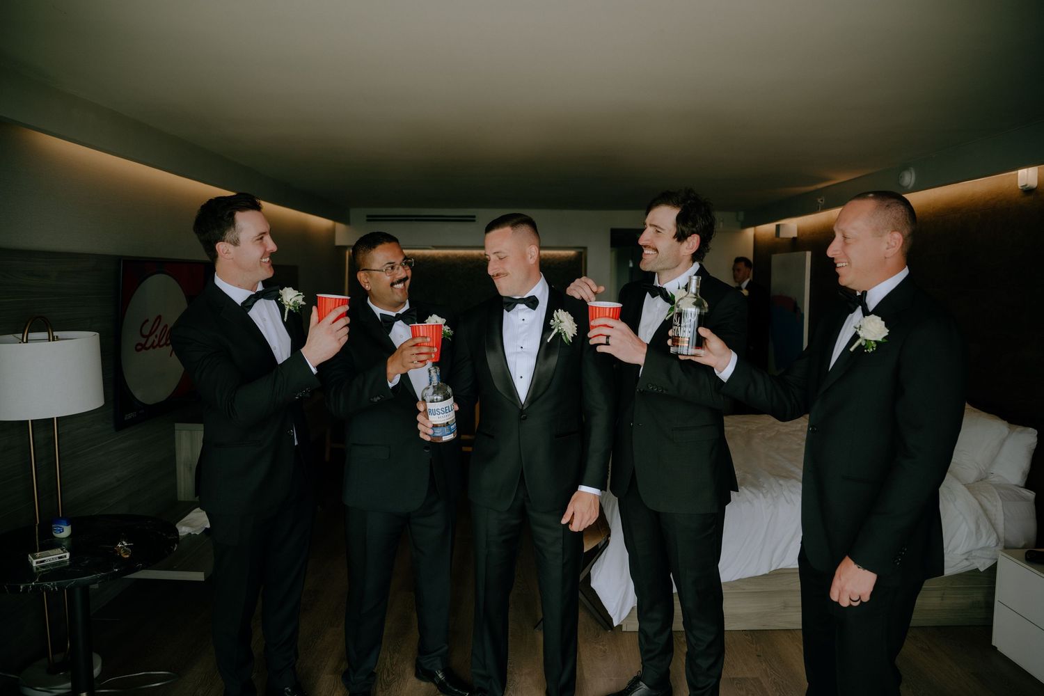 Groomsmen in black tuxedos toasting with red cups in a hotel room before the wedding ceremony.