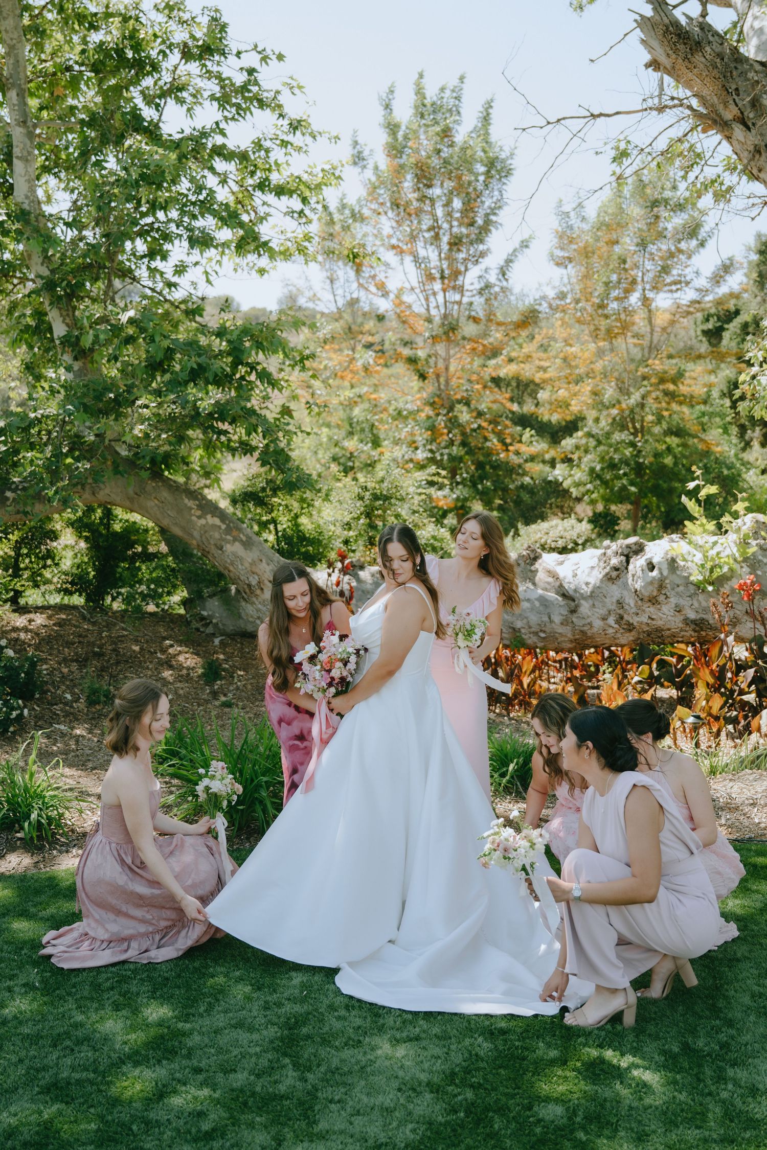 Bride in white gown surrounded by bridesmaids in blush pink dresses holding bouquets in a lush garden setting.