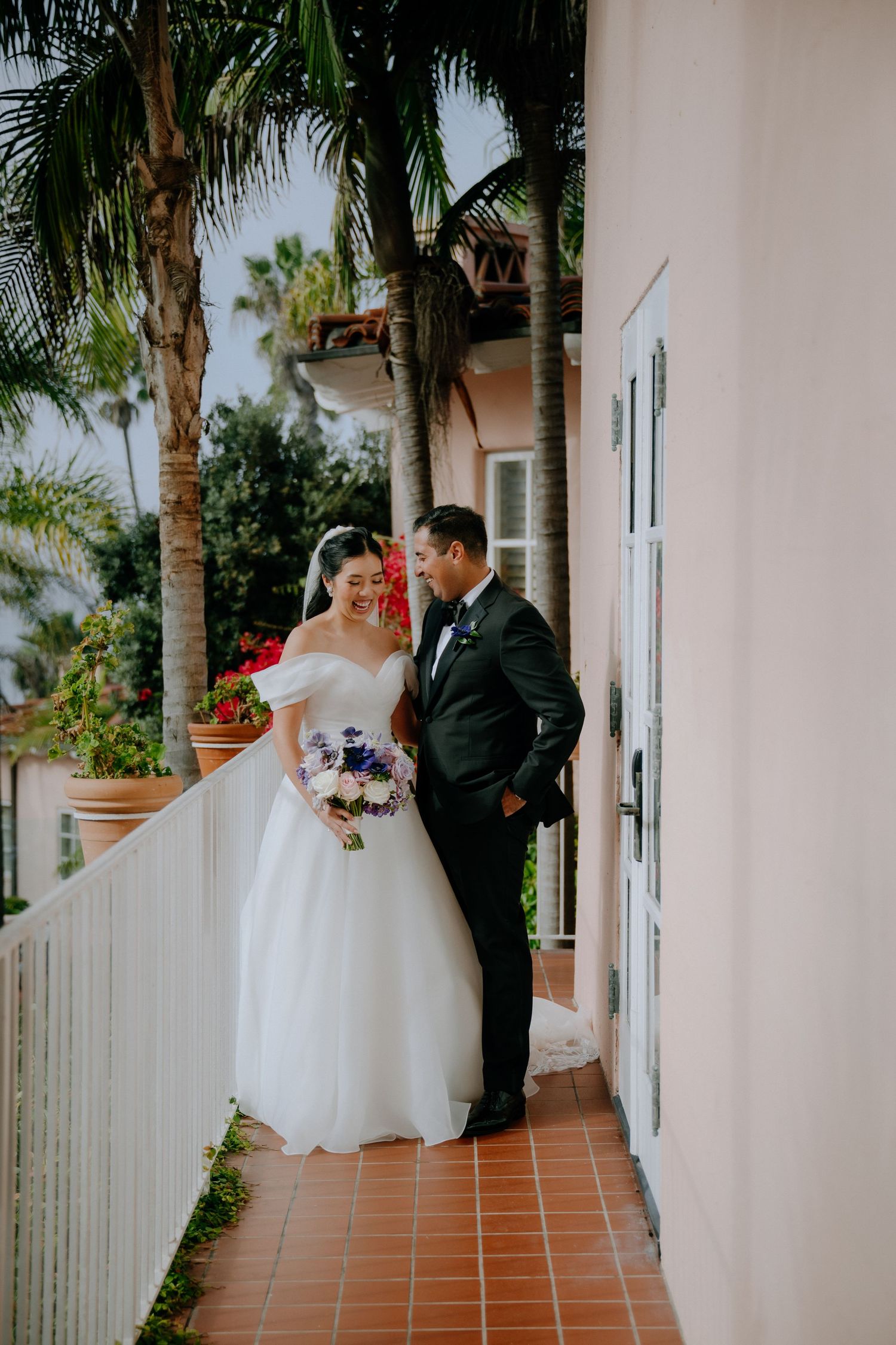 Bride in off-shoulder gown holding purple bouquet stands with groom in black tuxedo on balcony with palm trees.