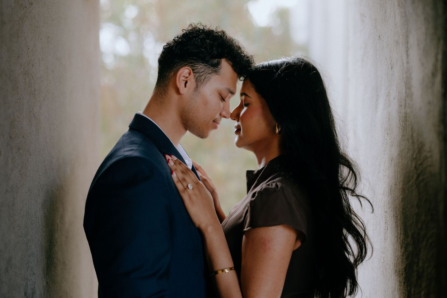A romantic couple shares an intimate moment, touching foreheads tenderly between stone columns in soft natural light.