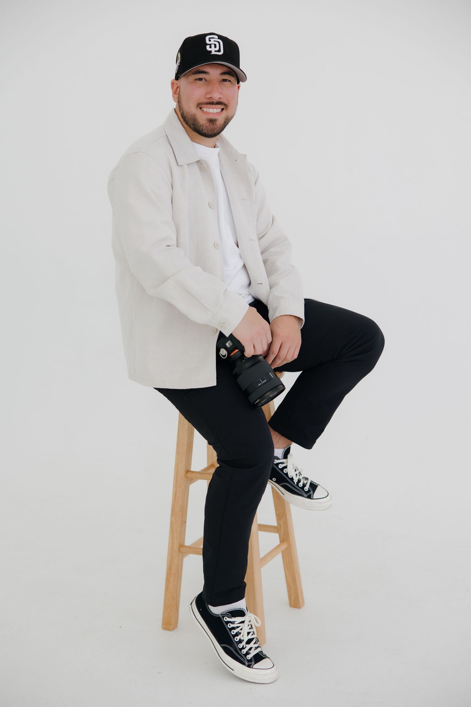 Smiling male wedding photographer sits on stool holding camera, wearing white jacket and Padres cap.