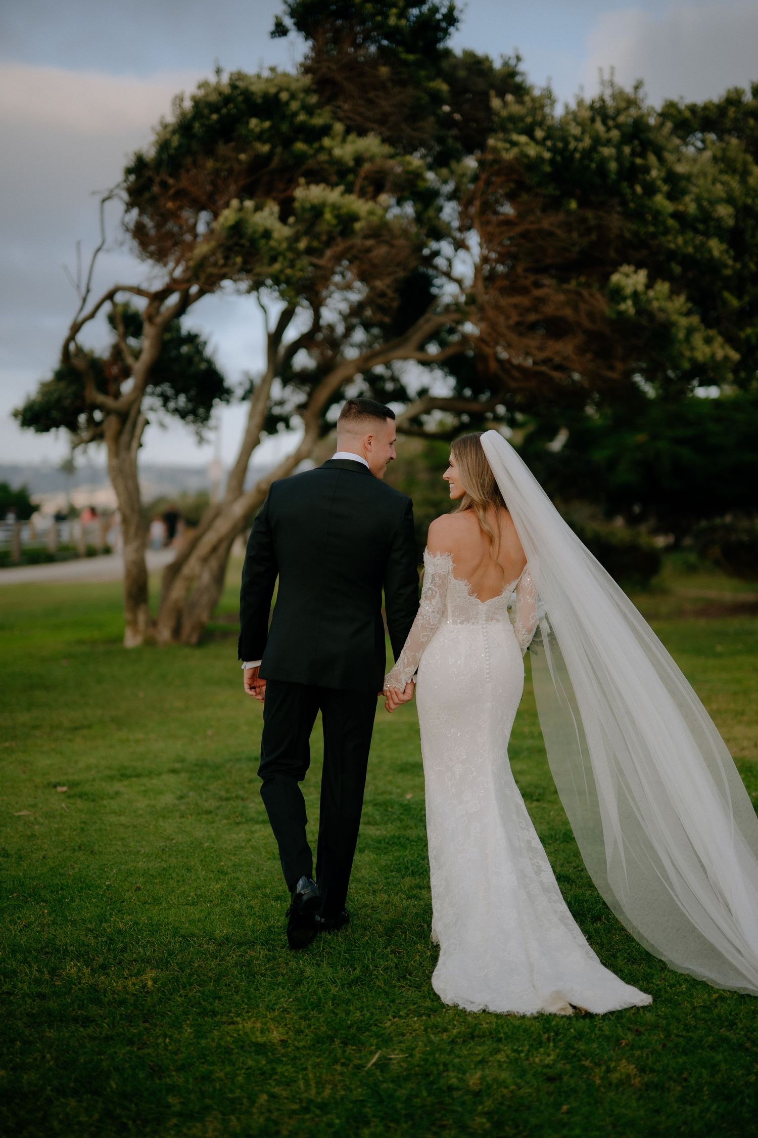 Bride and groom walking hand in hand on green lawn, bride in lace gown with flowing veil, groom in black suit.