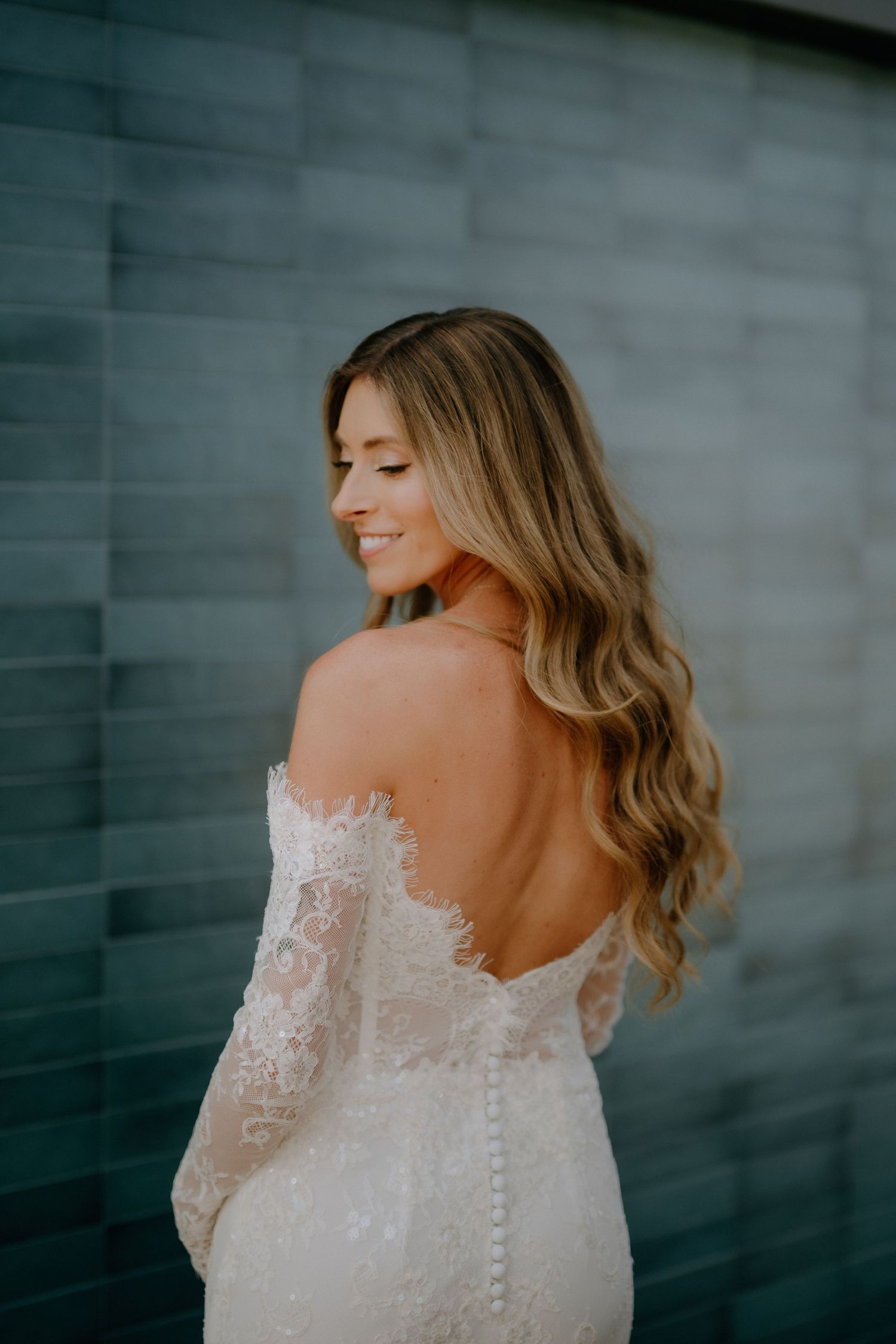 Bride in a backless lace off-shoulder wedding gown smiling over her shoulder against a tiled wall.