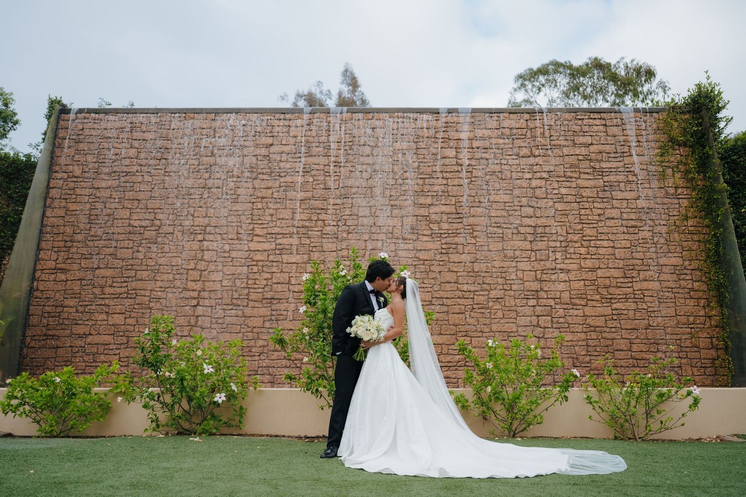 Bride and groom share a romantic kiss in front of a stunning brick waterfall wall surrounded by lush greenery.