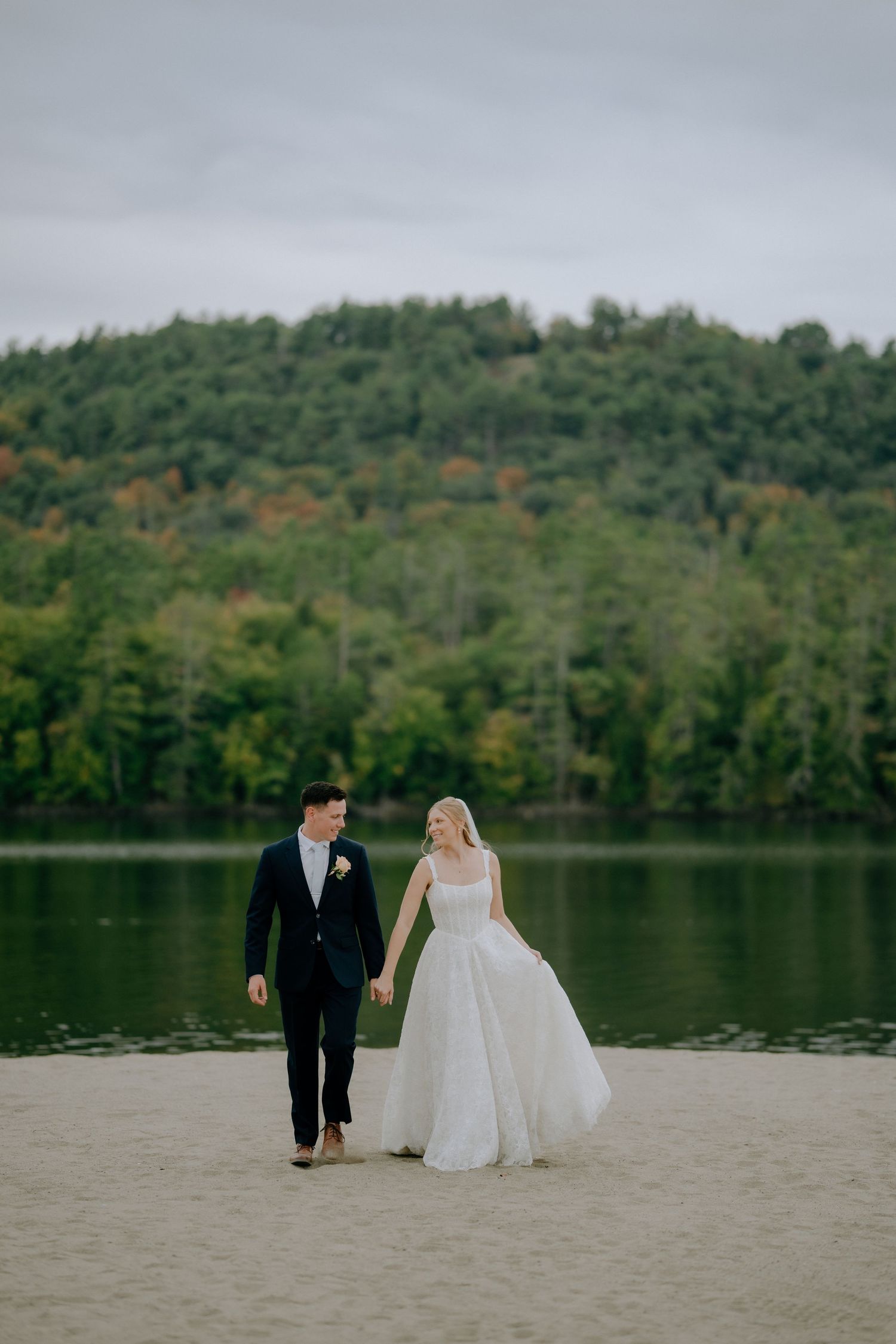 Bride and groom walk hand-in-hand on sandy beach with lush green forest and calm lake backdrop.