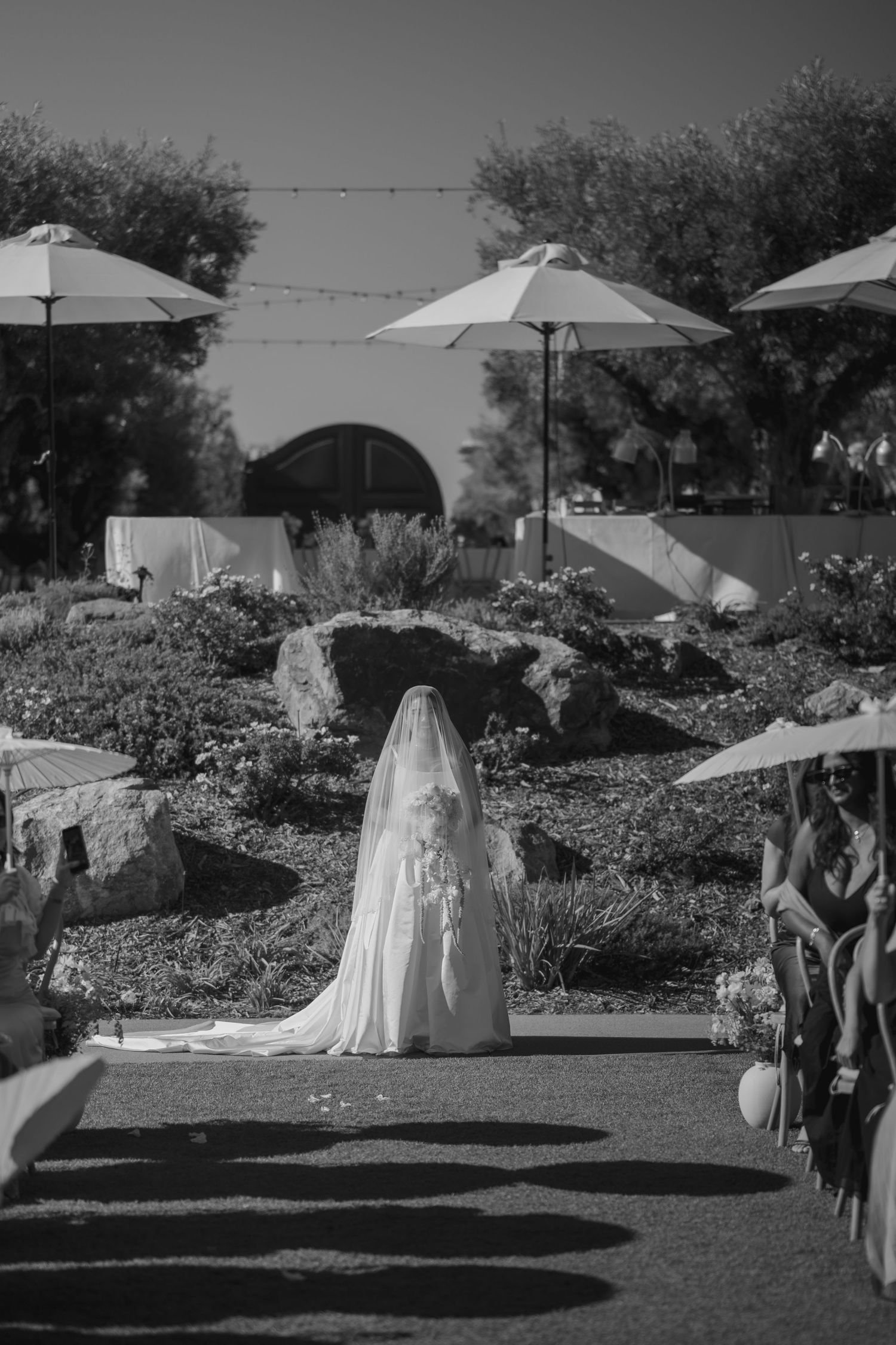 Black and white photo of a bride in a white gown and veil walking down an outdoor aisle at a garden wedding.