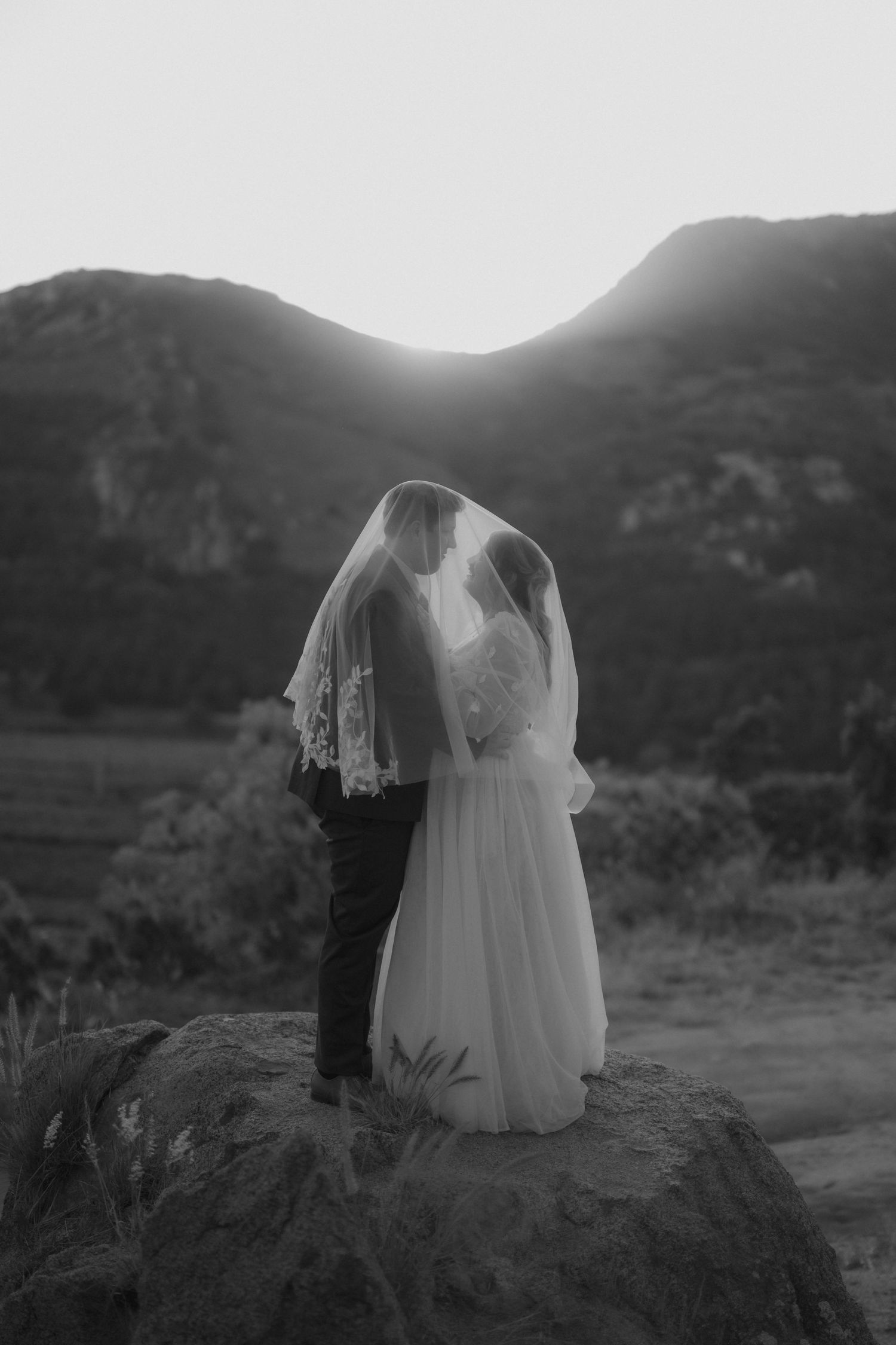 Black and white photo of bride and groom kissing under veil on rock with mountain sunset backdrop.