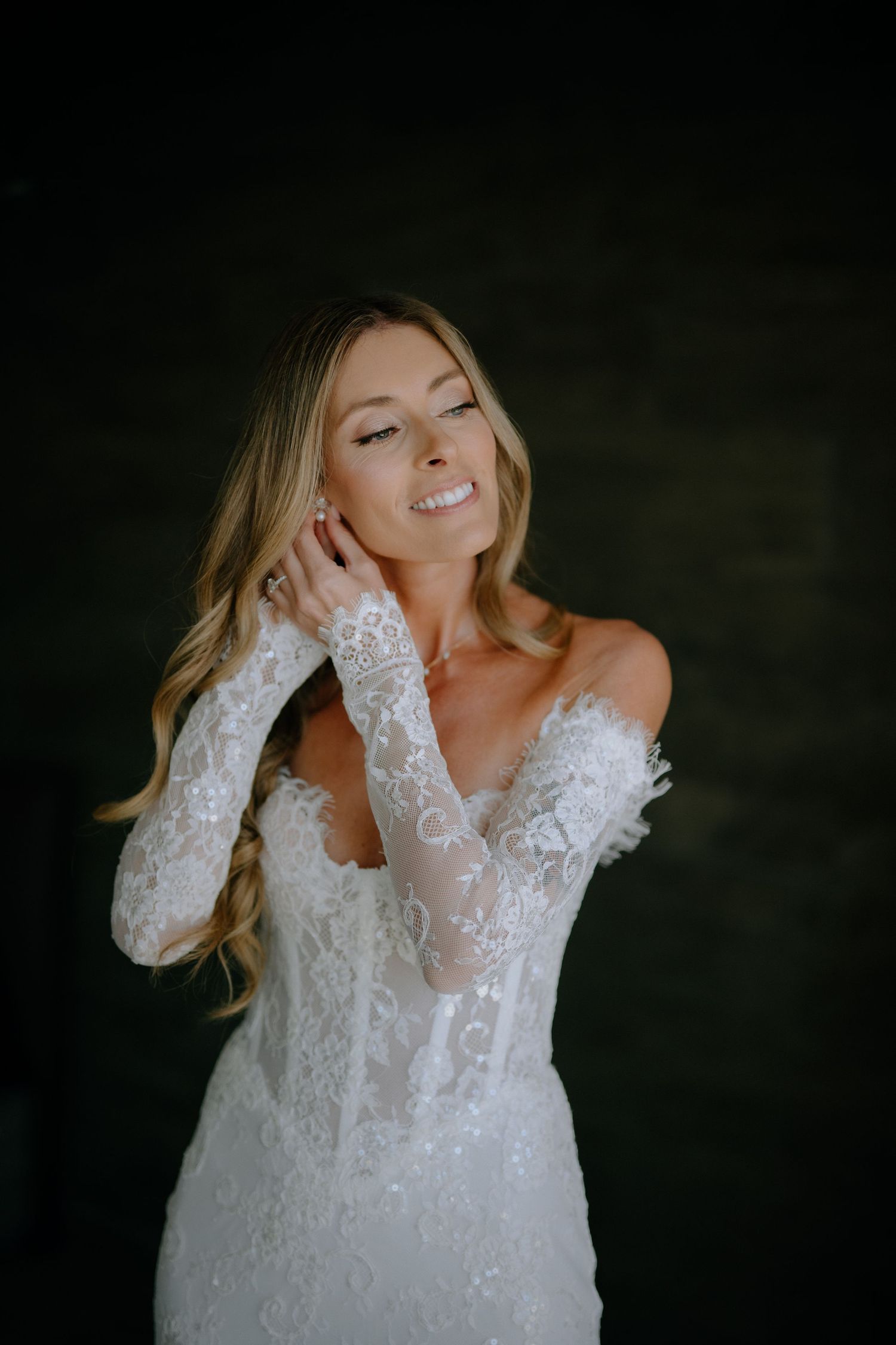 Elegant bride in off-shoulder lace wedding gown with long sleeves smiles against dark background.