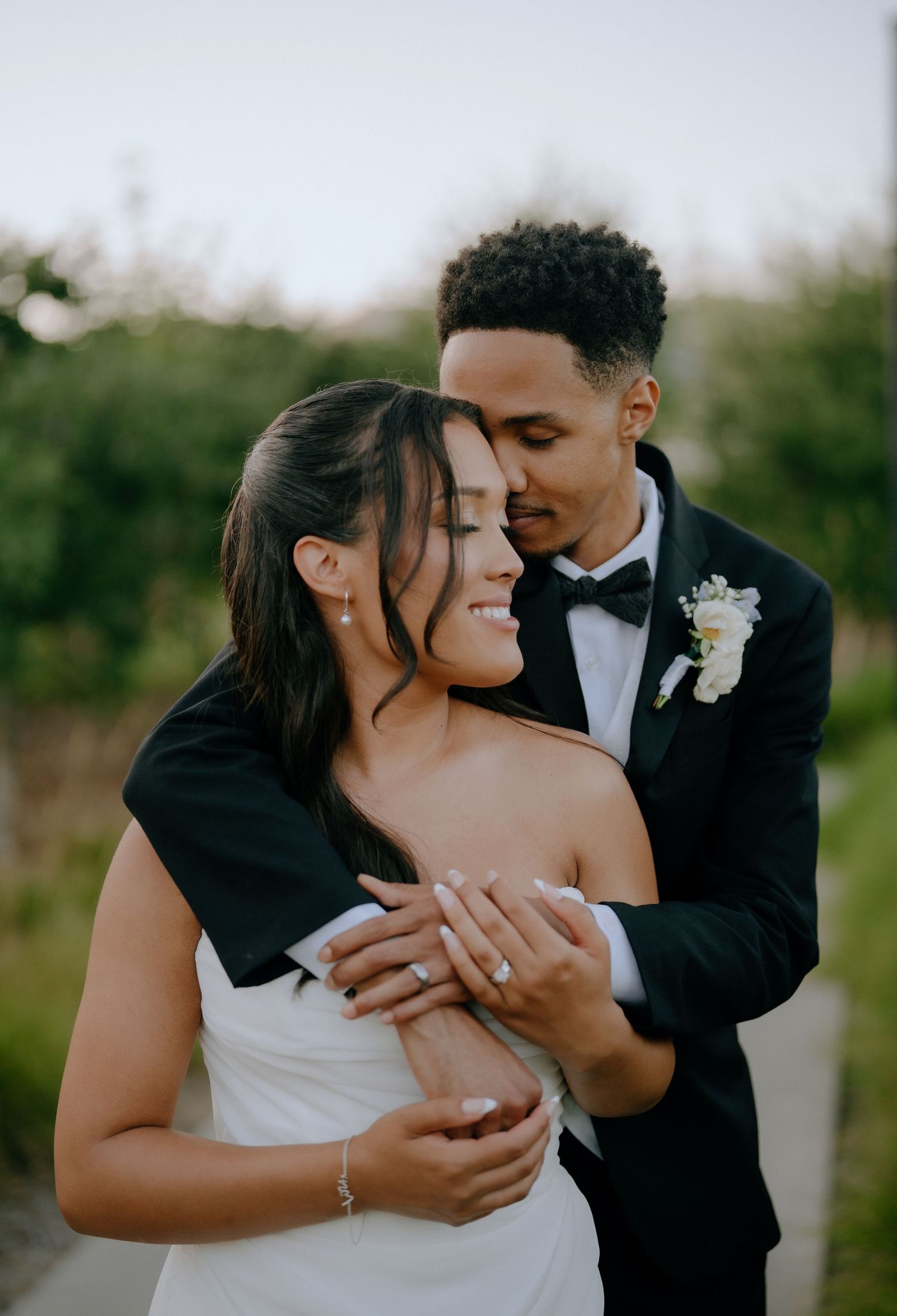 A bride and groom share a tender embrace outdoors, with the groom in a tuxedo holding the smiling bride in a white gown.