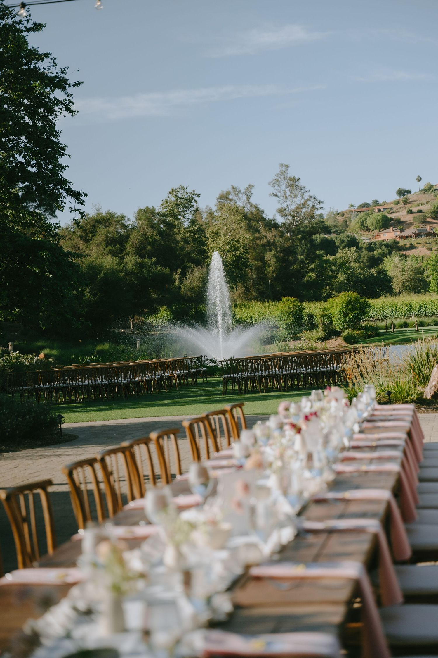 Elegant outdoor wedding reception with long dining tables, floral centerpieces, and a fountain surrounded by lush greenery.