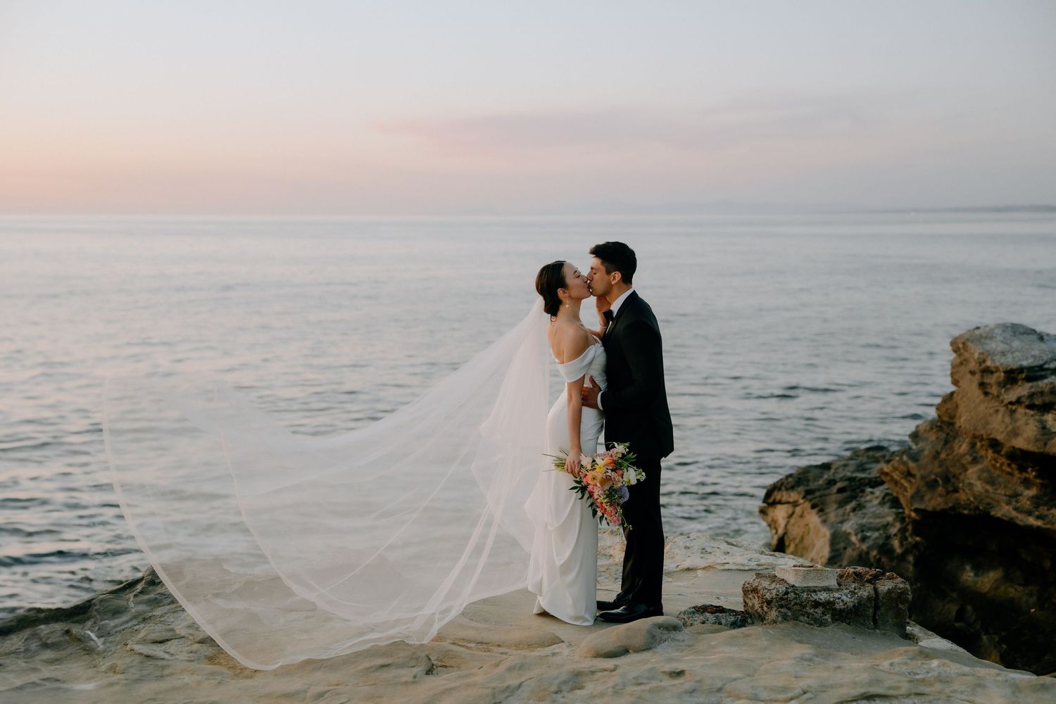 Bride and groom kissing on rocky oceanside cliff at sunset, wedding veil flowing in breeze.