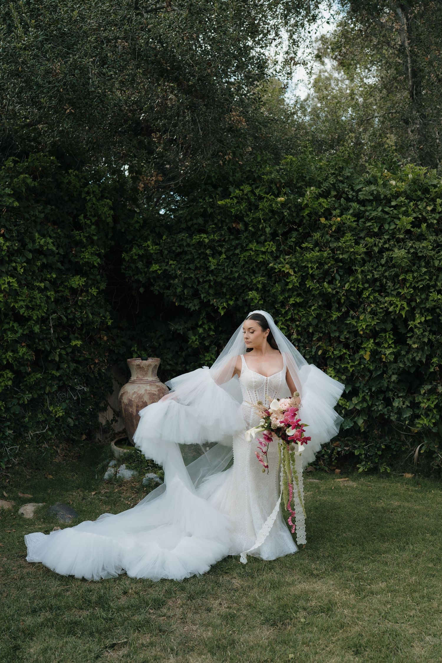 Bride in fitted white gown with dramatic tulle veil holds colorful bouquet in lush garden setting.