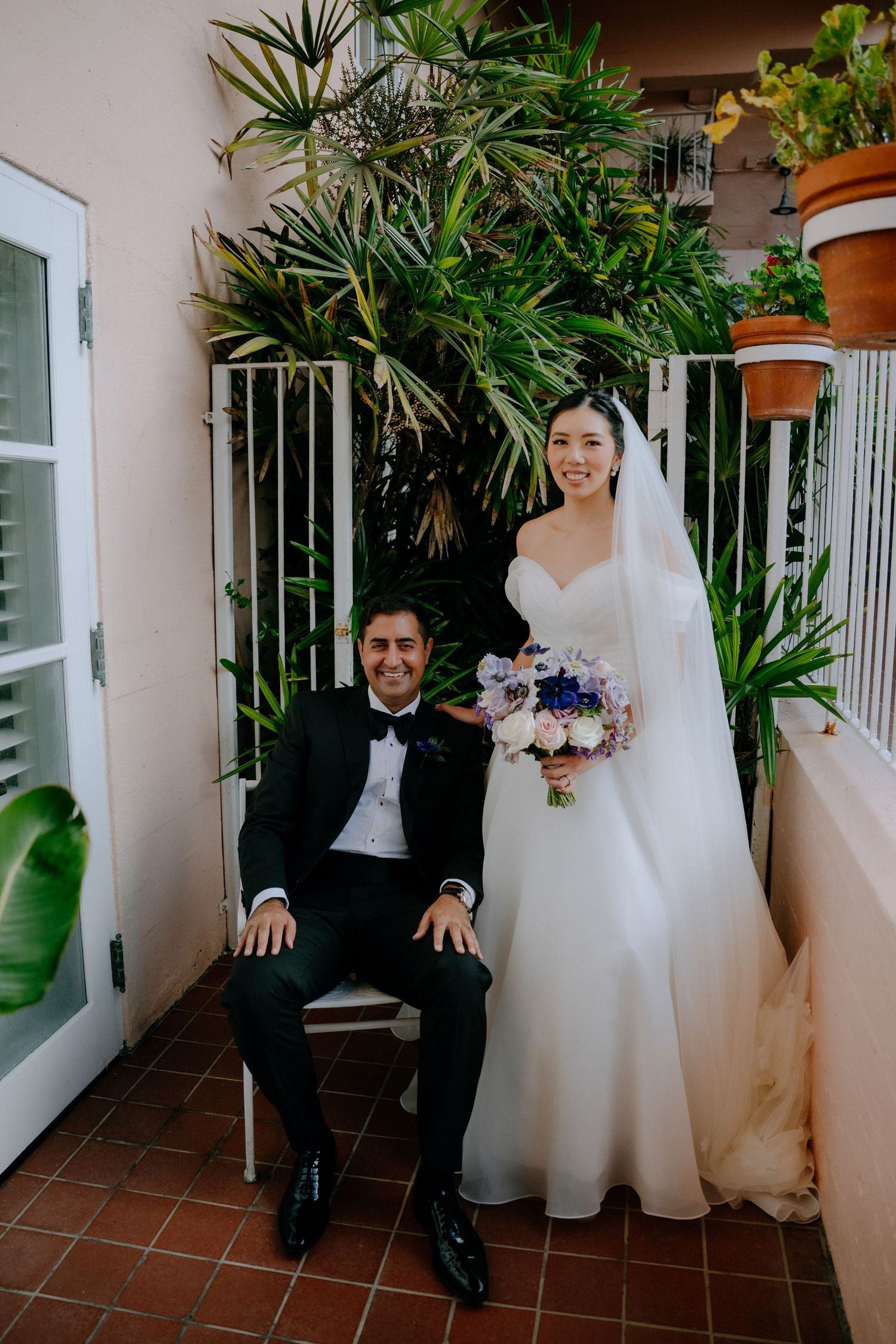 Bride in white gown holding bouquet stands beside groom in tuxedo seated on balcony with tropical plants.