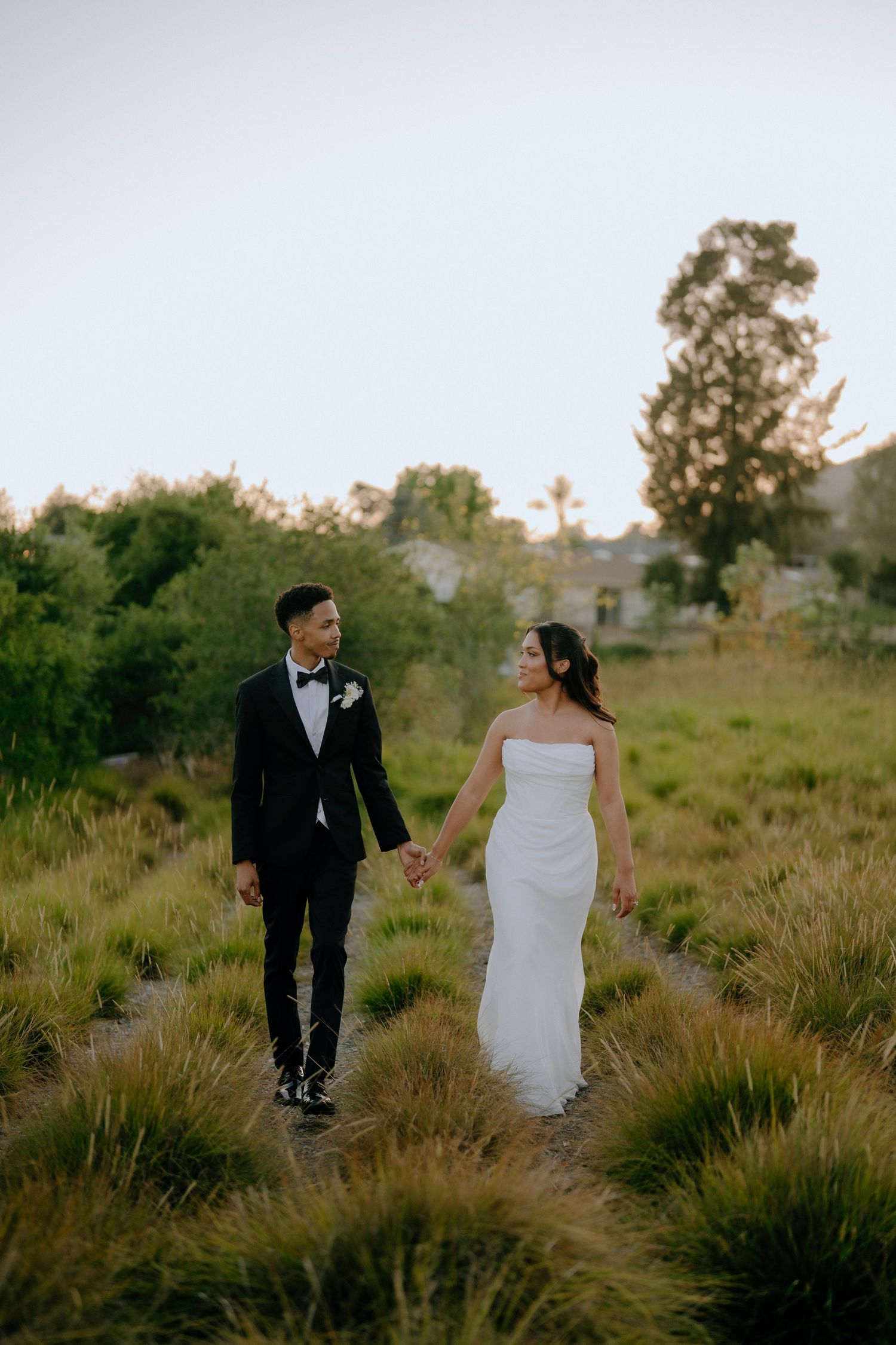 A couple in wedding attire walks hand-in-hand through a lush green garden path at dusk.