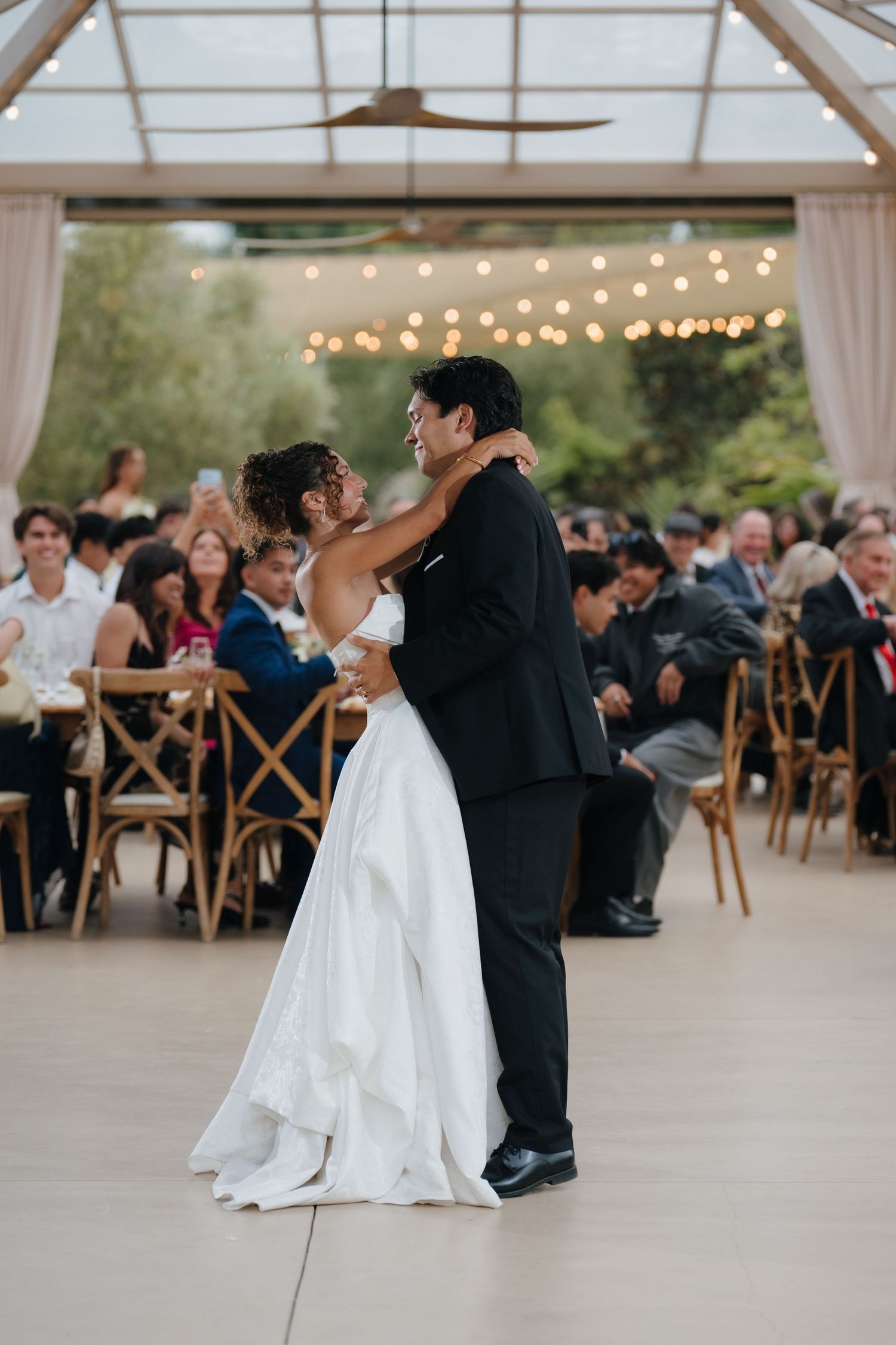 A bride and groom share their first dance under string lights at an elegant outdoor wedding reception with guests seated nearby.