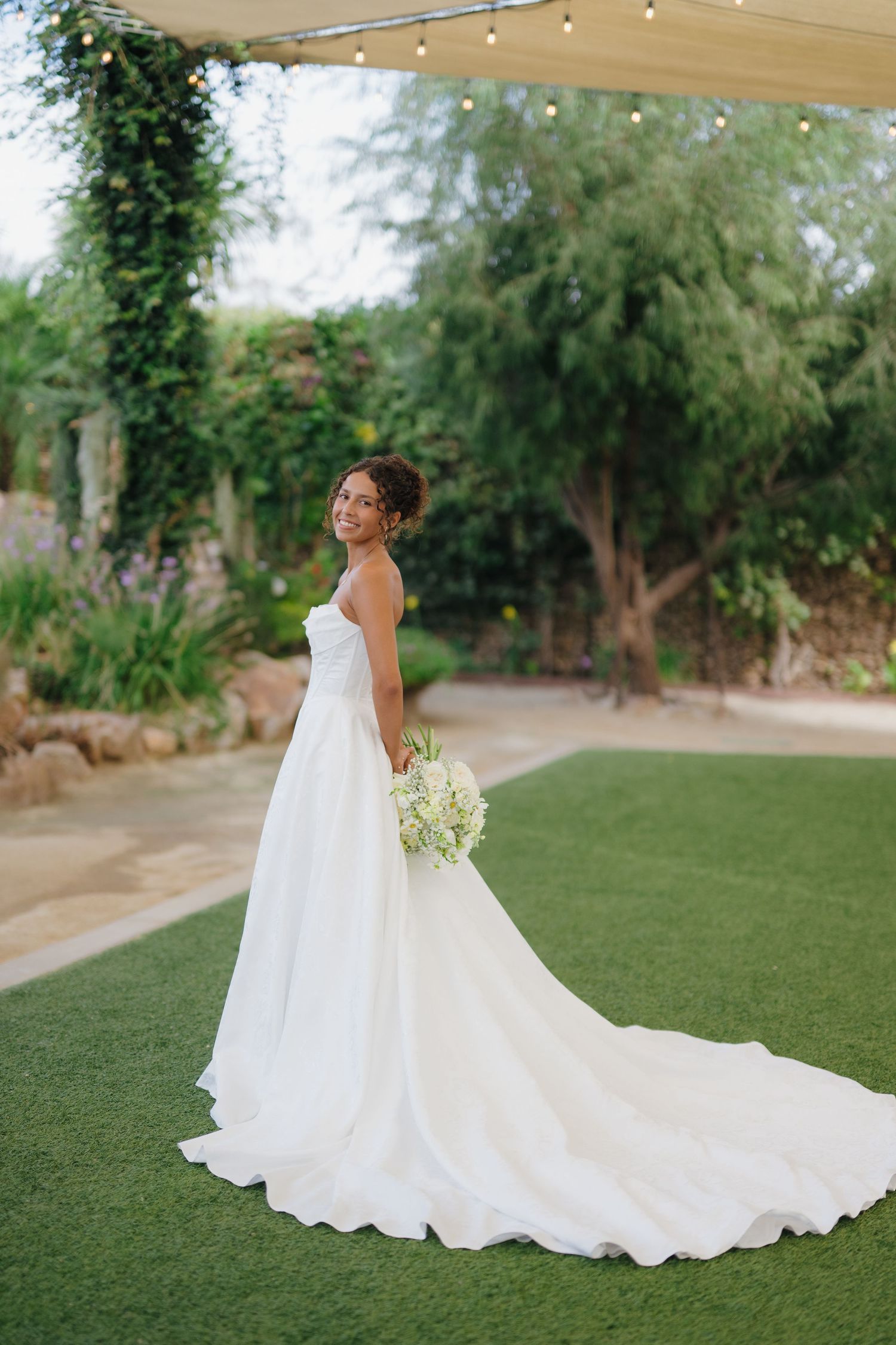 Smiling bride in strapless white ballgown with long train holds white bouquet in garden venue.