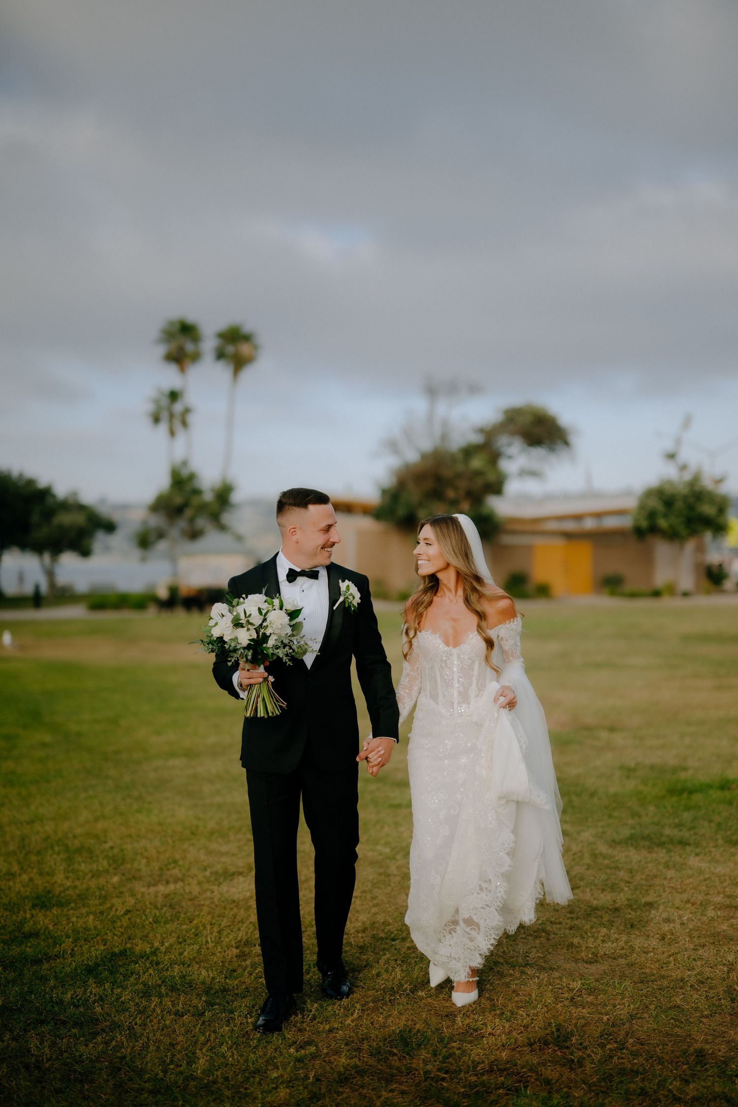 Bride and groom walking hand in hand on a green lawn with palm trees under a cloudy sky.